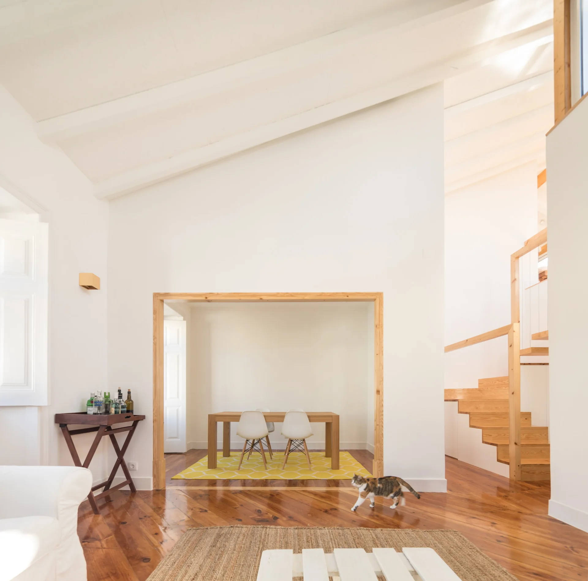 A bright white room features polished wood floors, a wooden staircase, and a wood-trimmed opening leading to a dining nook with a yellow geometric rug.