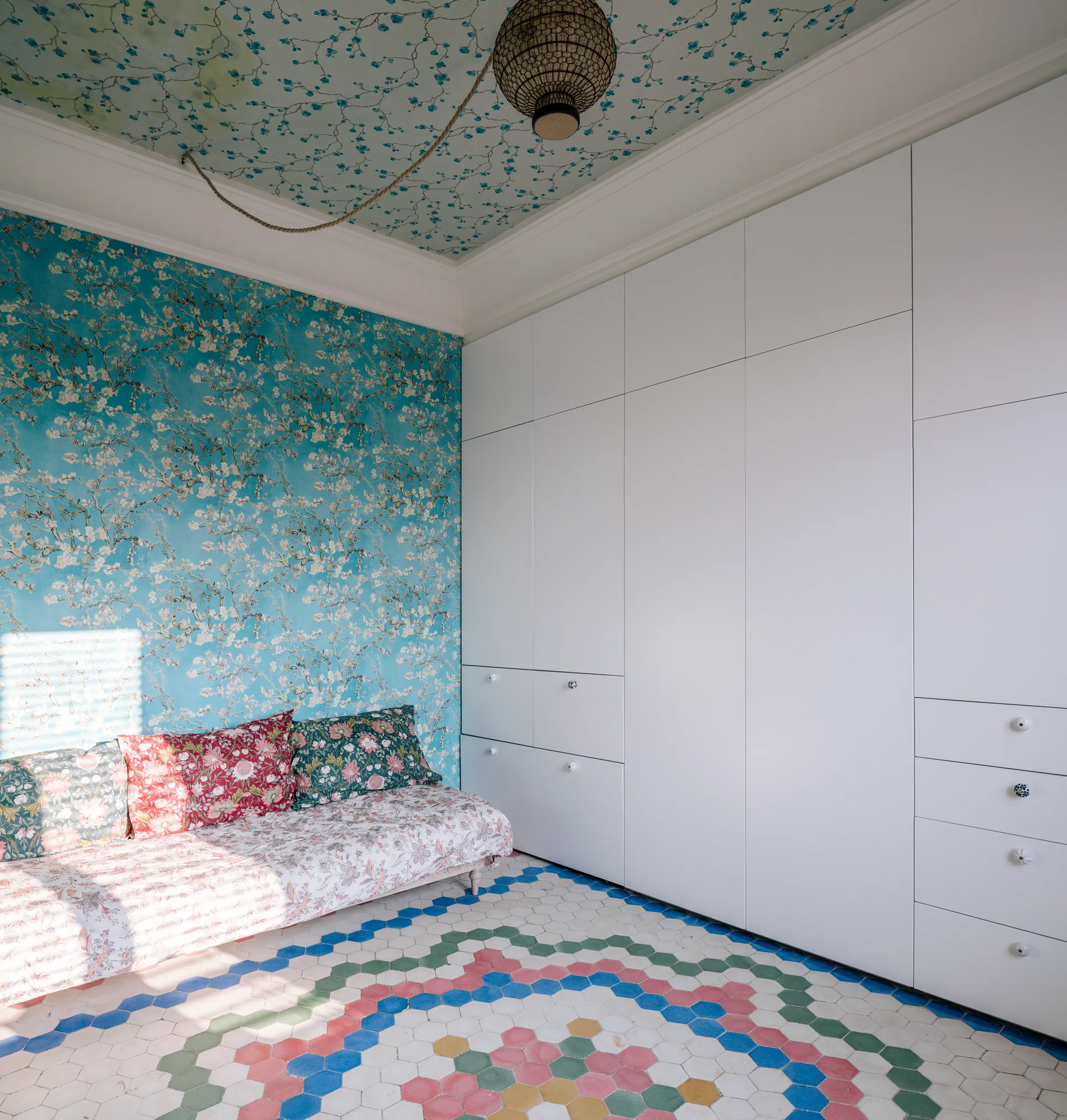 A bedroom featuring turquoise floral wallpaper on the wall and ceiling, a colorful patterned hexagon tile floor, and a large white built-in wardrobe.