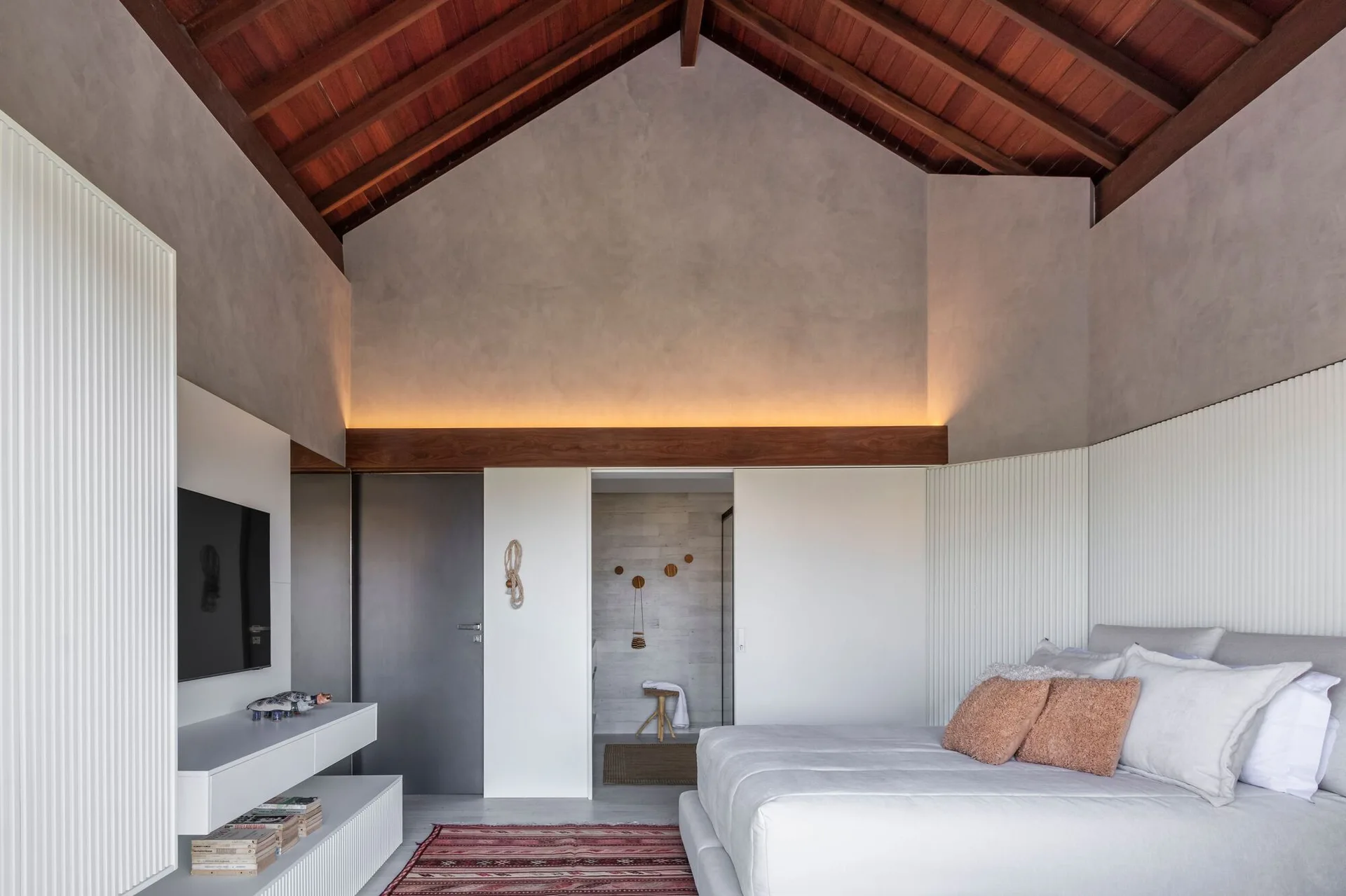 A minimalist bedroom featuring a high vaulted wood ceiling, white slatted wall paneling, a platform bed, and a wall-mounted television.