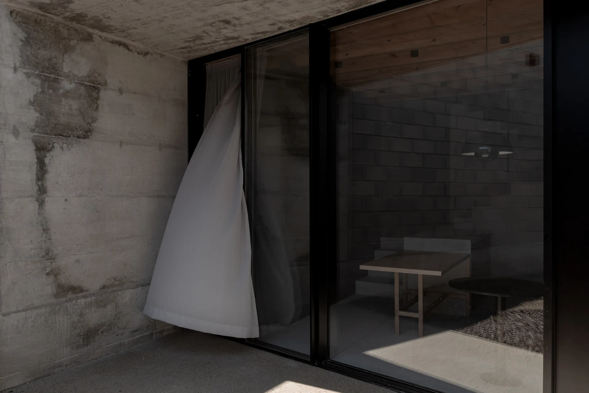 A view through a large glass sliding door reveals a minimalist dining room with a wooden table, concrete block walls, and a white breeze-blown curtain.
