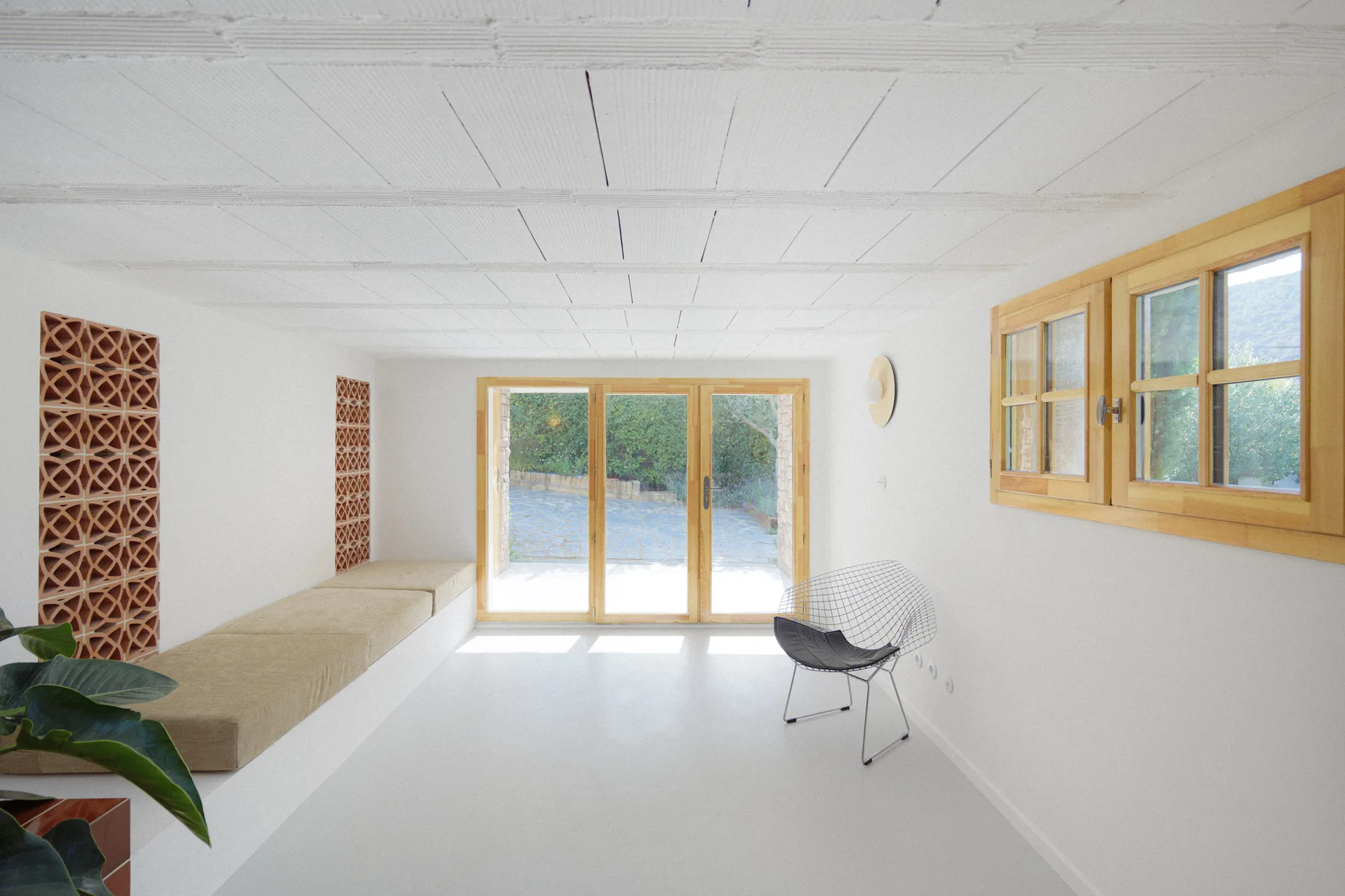A minimalist white sunroom features a long built-in bench with beige cushions, terracotta decorative screen blocks, and wood-framed glass doors.