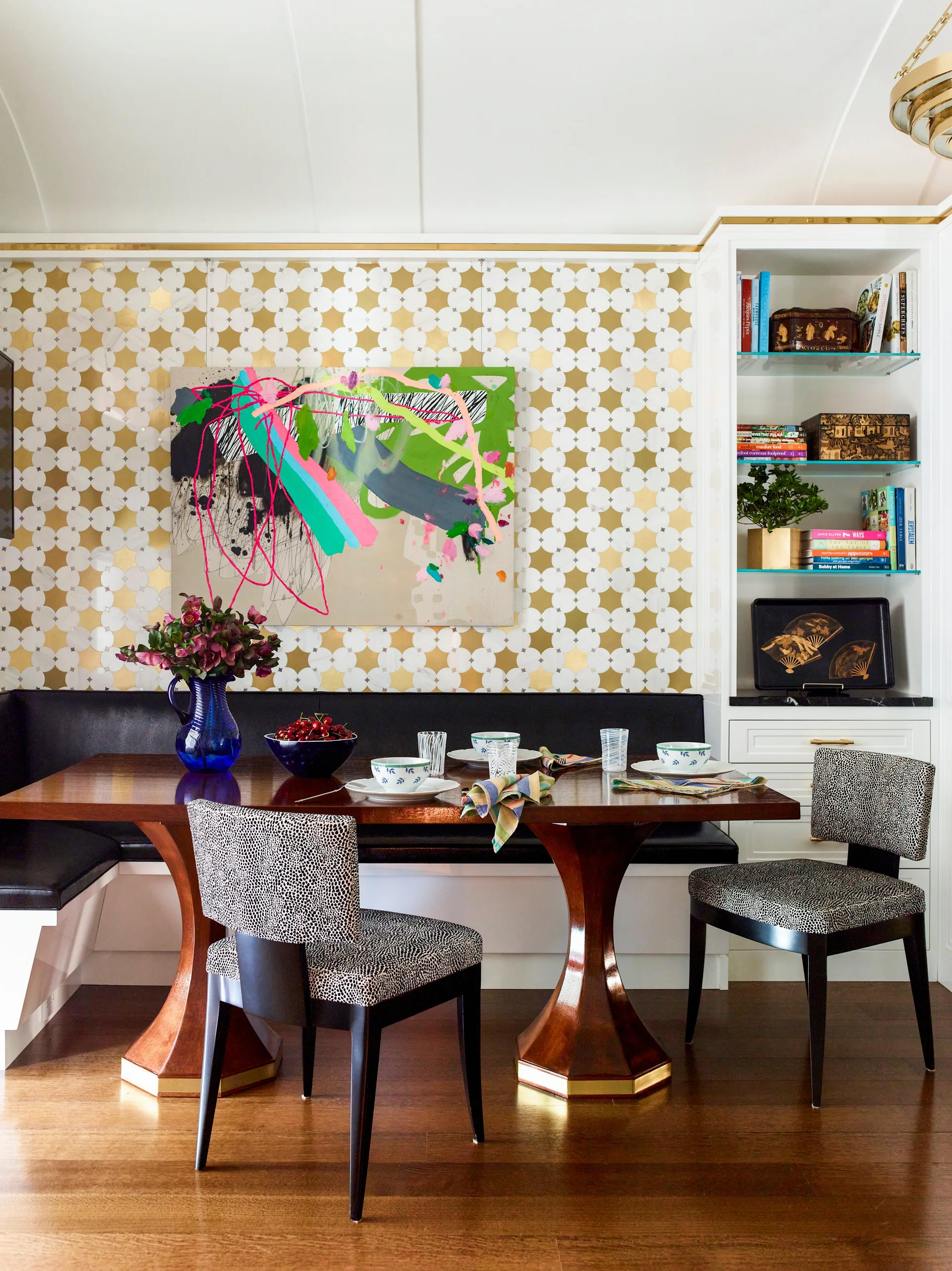 A dining nook features a wooden pedestal table surrounded by animal print chairs and a black banquette against gold star-patterned wallpaper.
