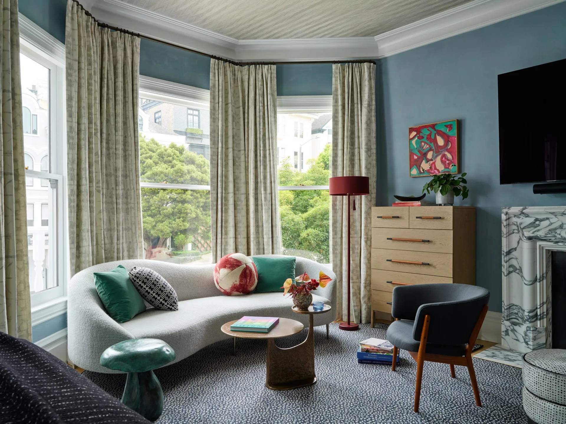 An eclectic living room with a curved grey sofa in a bay window, blue walls, a light wood dresser, and a marble fireplace.