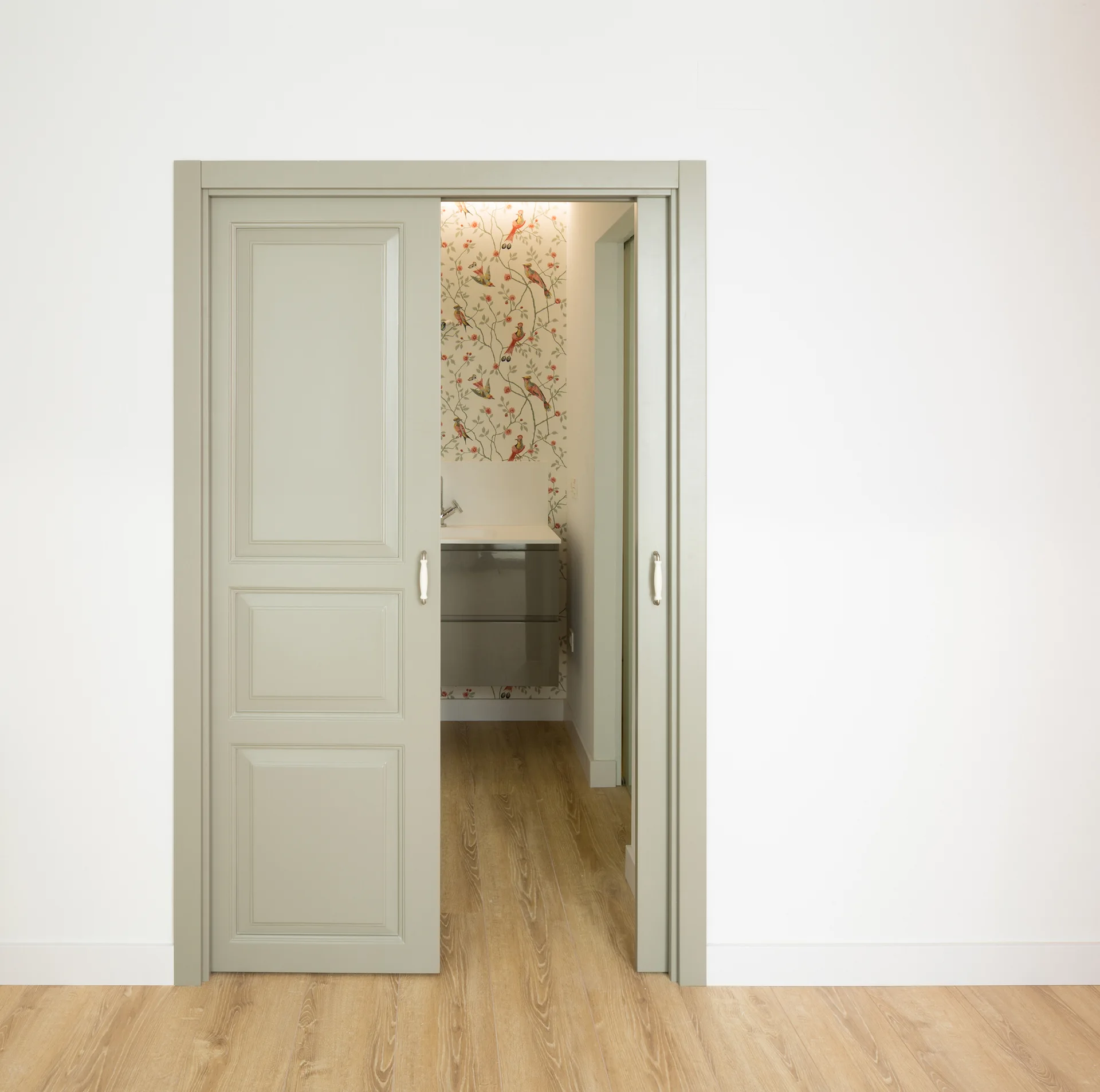 A pair of sage green paneled pocket doors open to reveal a bathroom with bird-patterned wallpaper and a grey floating vanity over wood floors.