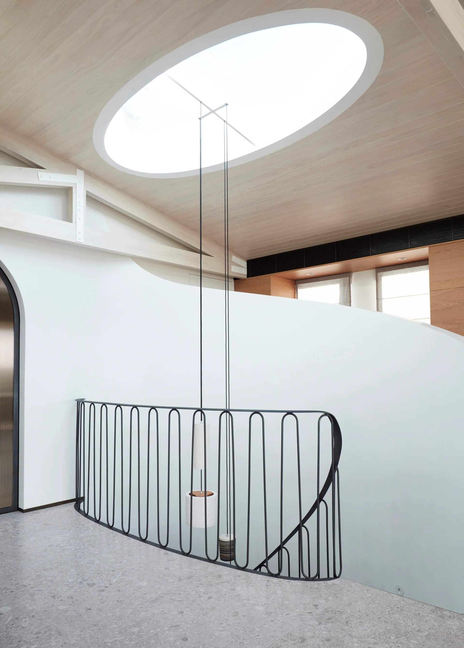 A modern stairwell features a curved black metal railing, speckled grey flooring, and a large oval skylight set into a light wood ceiling.