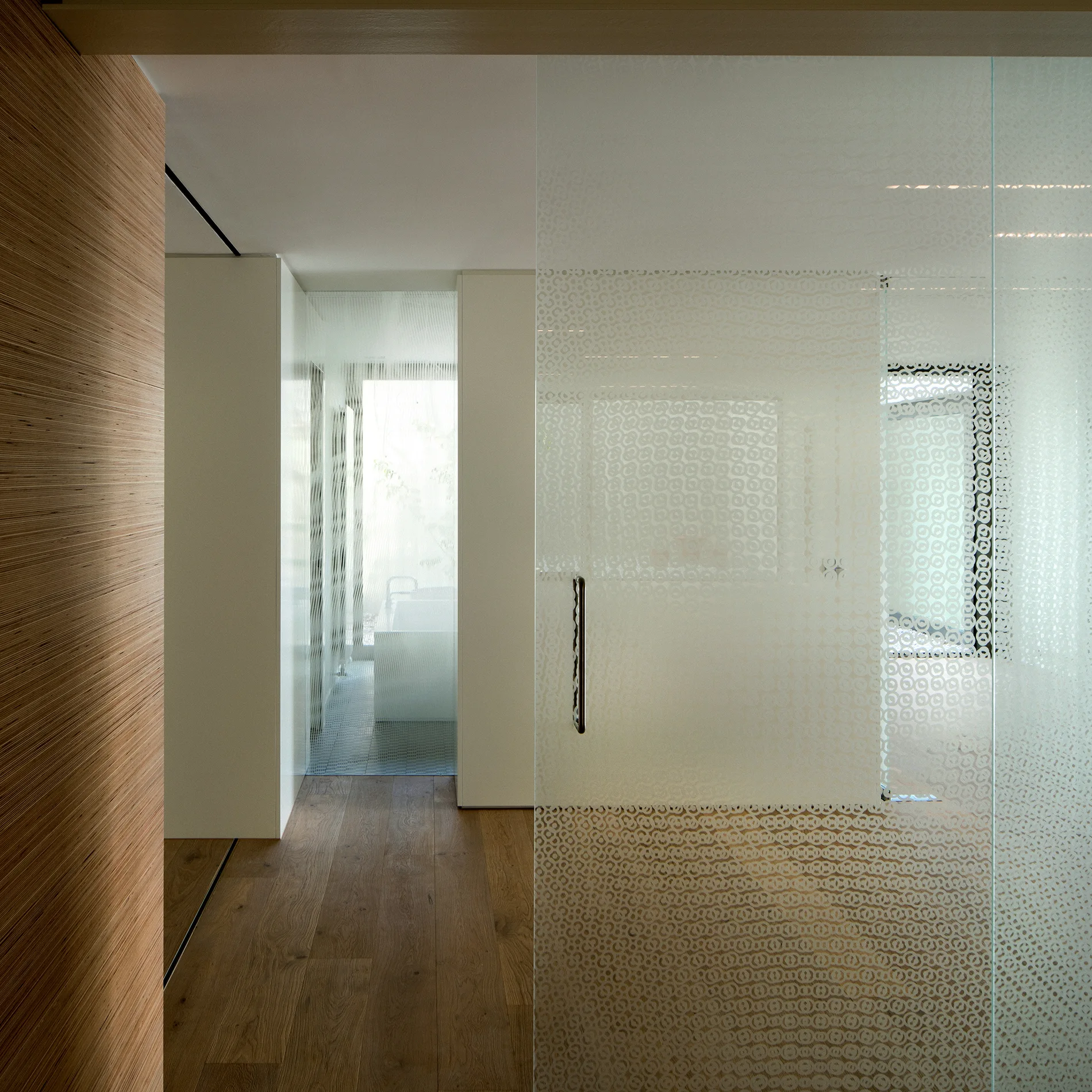 A modern hallway with wood plank flooring and textured wood wall paneling features a large sliding frosted glass partition leading to a bright bathroom.