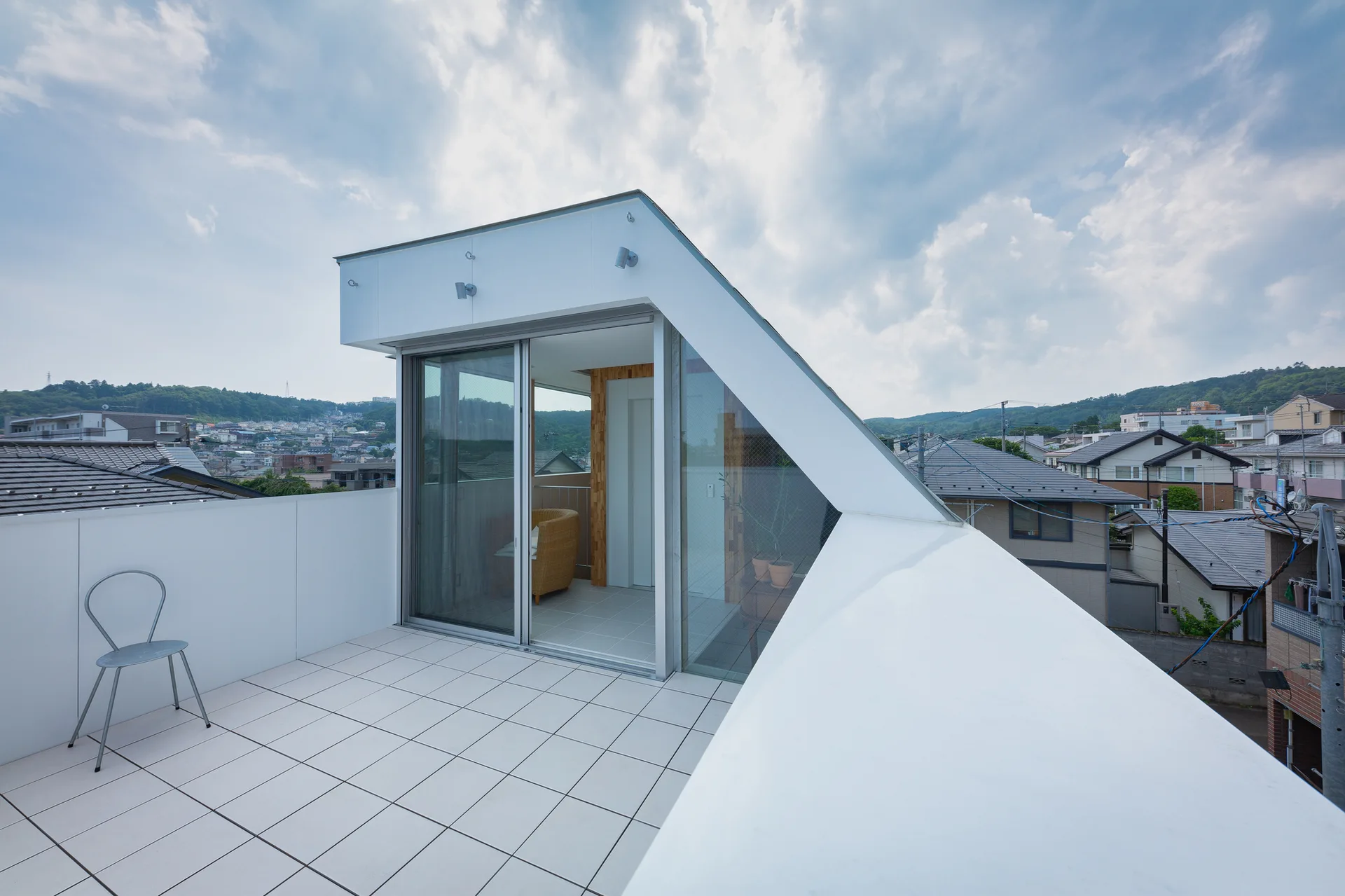 A modern white tiled rooftop terrace features a metal chair and a sloped architectural structure with glass sliding doors overlooking a neighborhood.