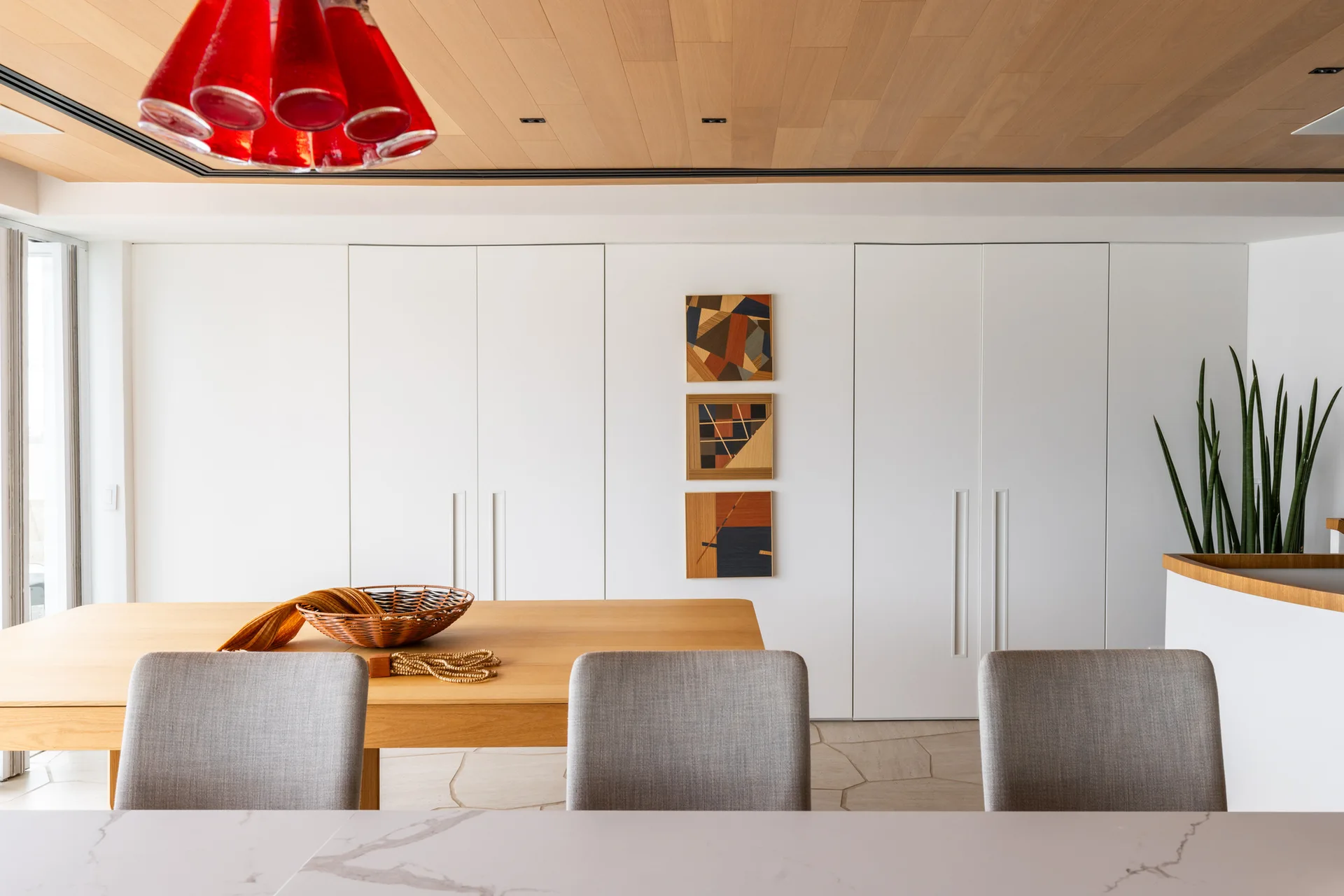 A modern dining room features a light wood ceiling, flat-panel white cabinetry, a wooden table with gray upholstered chairs, and a red glass pendant light.