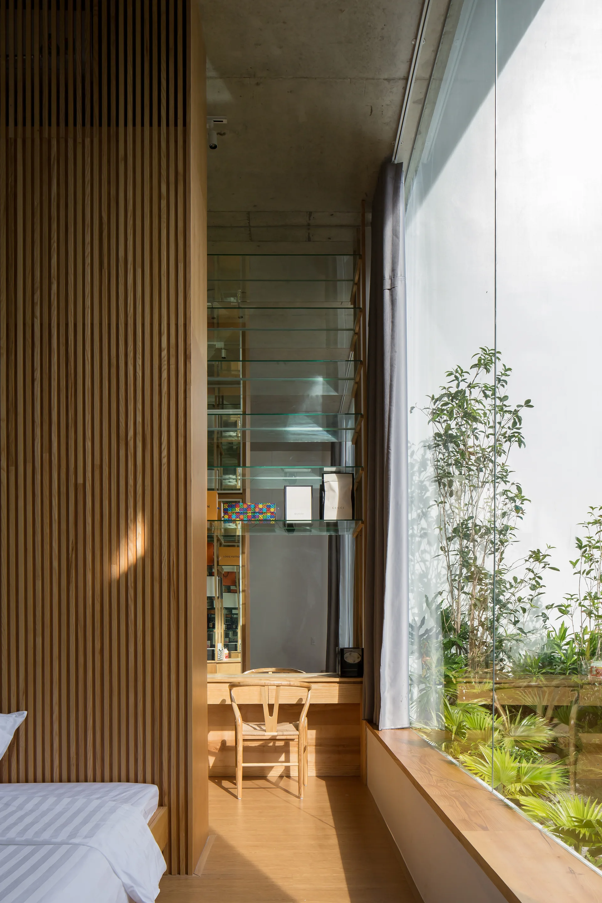 A modern bedroom features a vertical wood slat wall, a built-in wooden desk with glass shelving, and a glass window overlooking an outdoor garden.