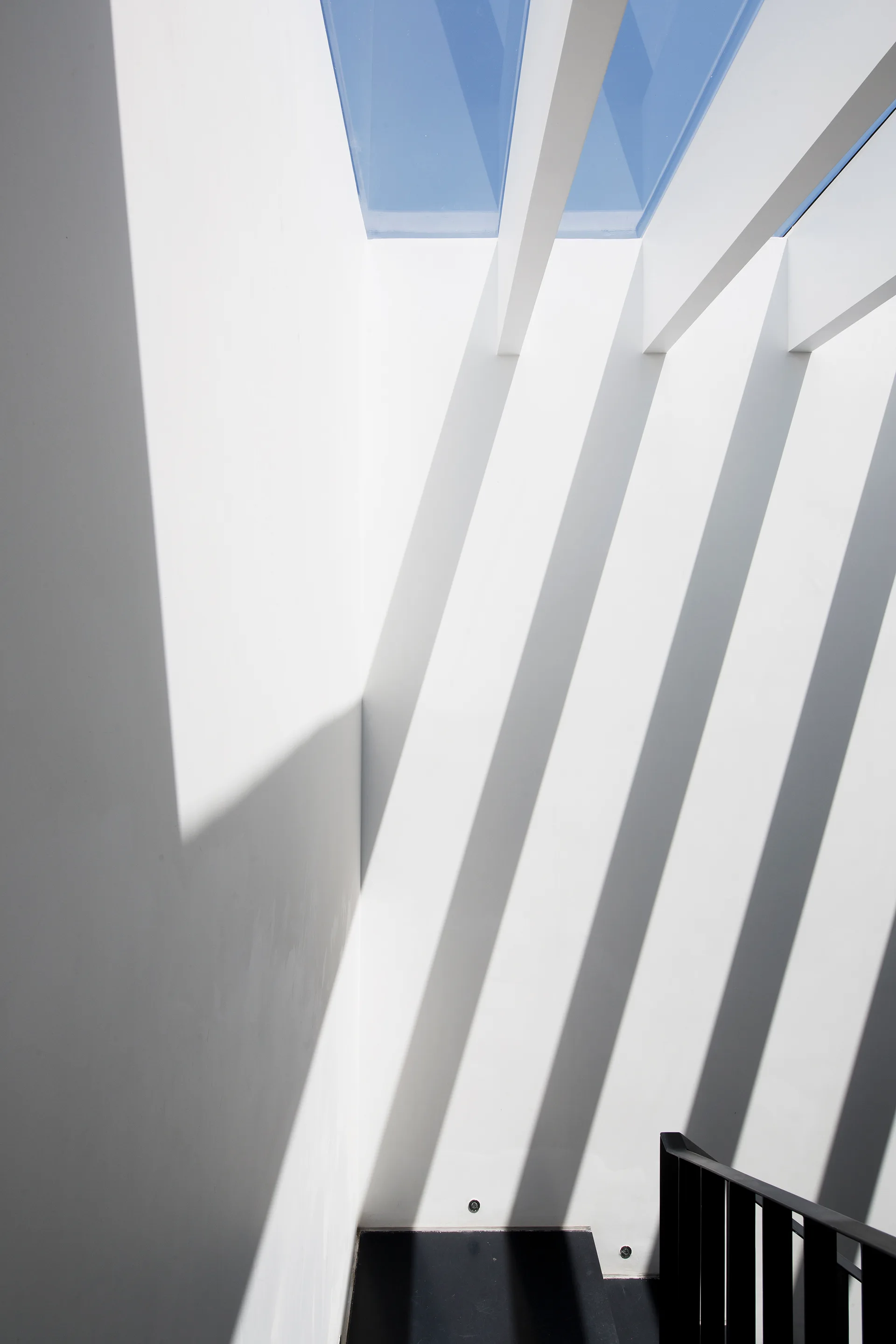 A minimalist white stairwell features a skylight with overhead beams that cast sharp parallel shadows along the tall plaster walls.