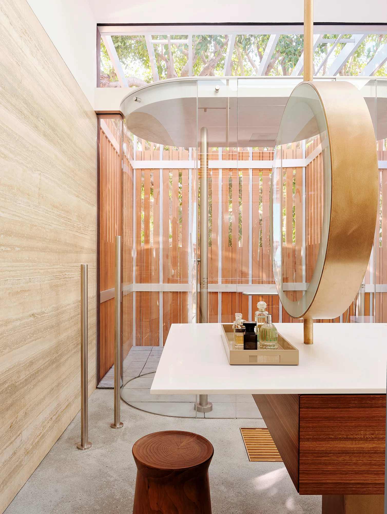 A modern bathroom features a round backlit mirror on a white countertop vanity, next to a travertine wall and glass shower with wood slat walls.