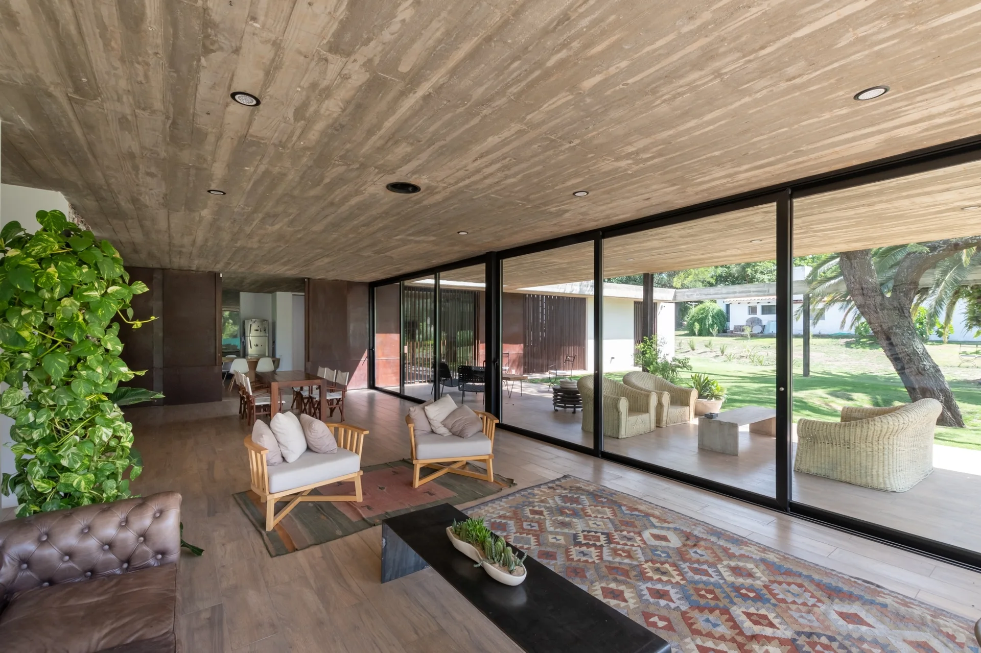 A contemporary living room featuring a board-formed concrete ceiling, wood plank floors, mid-century wood-framed armchairs, and sliding glass doors.