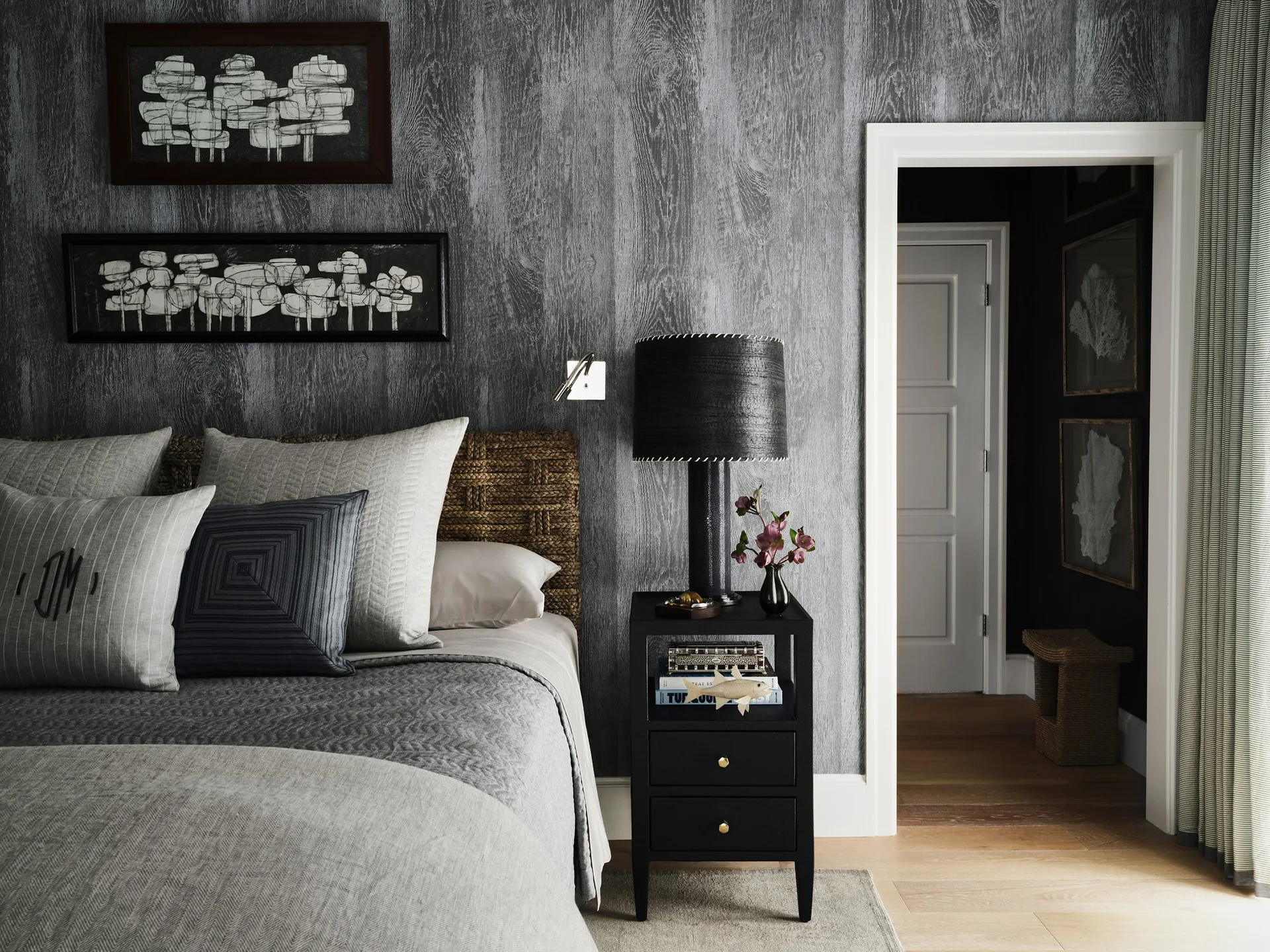 A bedroom wall covered in charcoal wood-grain wallpaper featuring a woven brown headboard, black nightstand, and doorway.