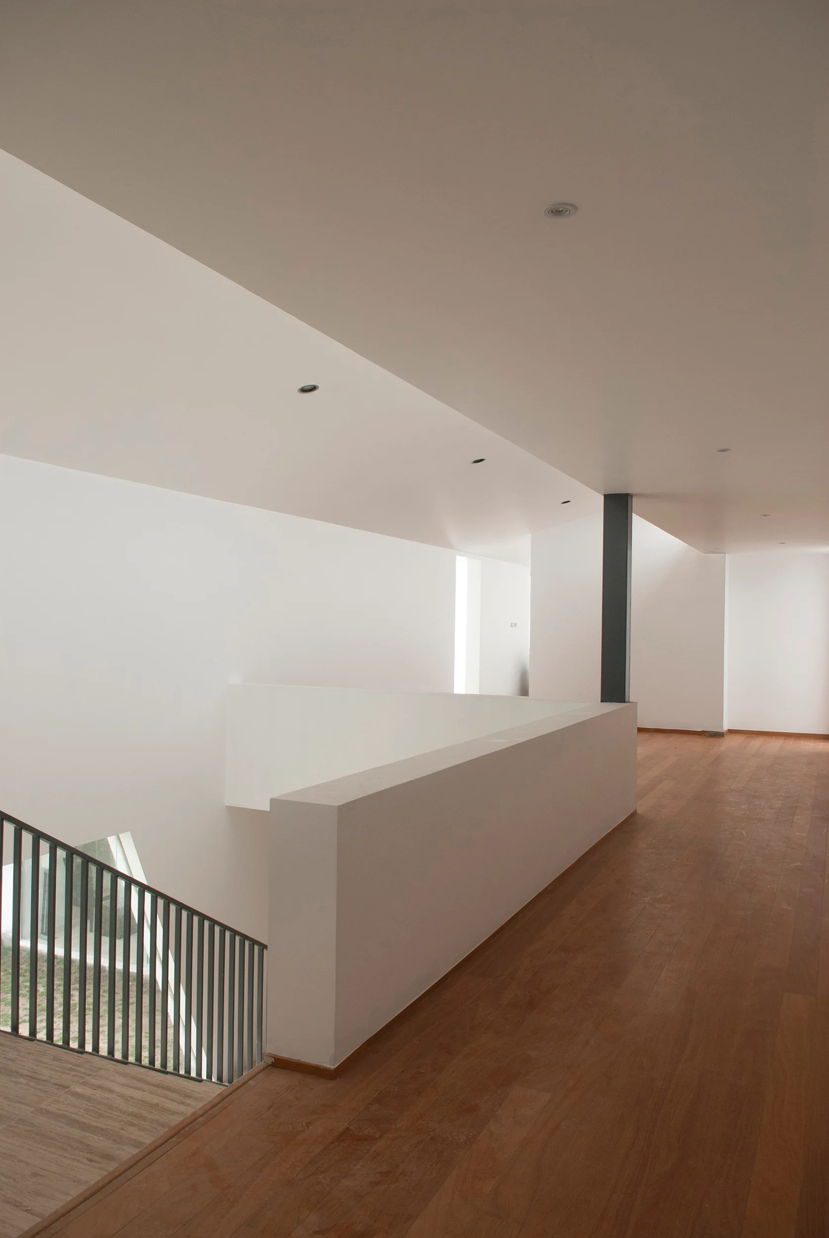 A minimalist interior landing features warm wood flooring, white walls, a solid white partition wall, and black metal stair railings.