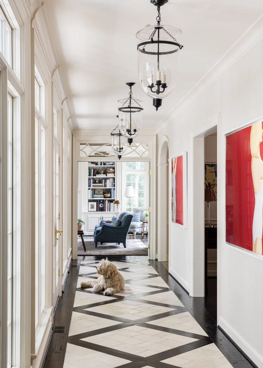 A long white hallway features three hanging glass lantern pendants, vibrant red wall art, and a geometric-patterned stone tile floor bordered by dark wood.