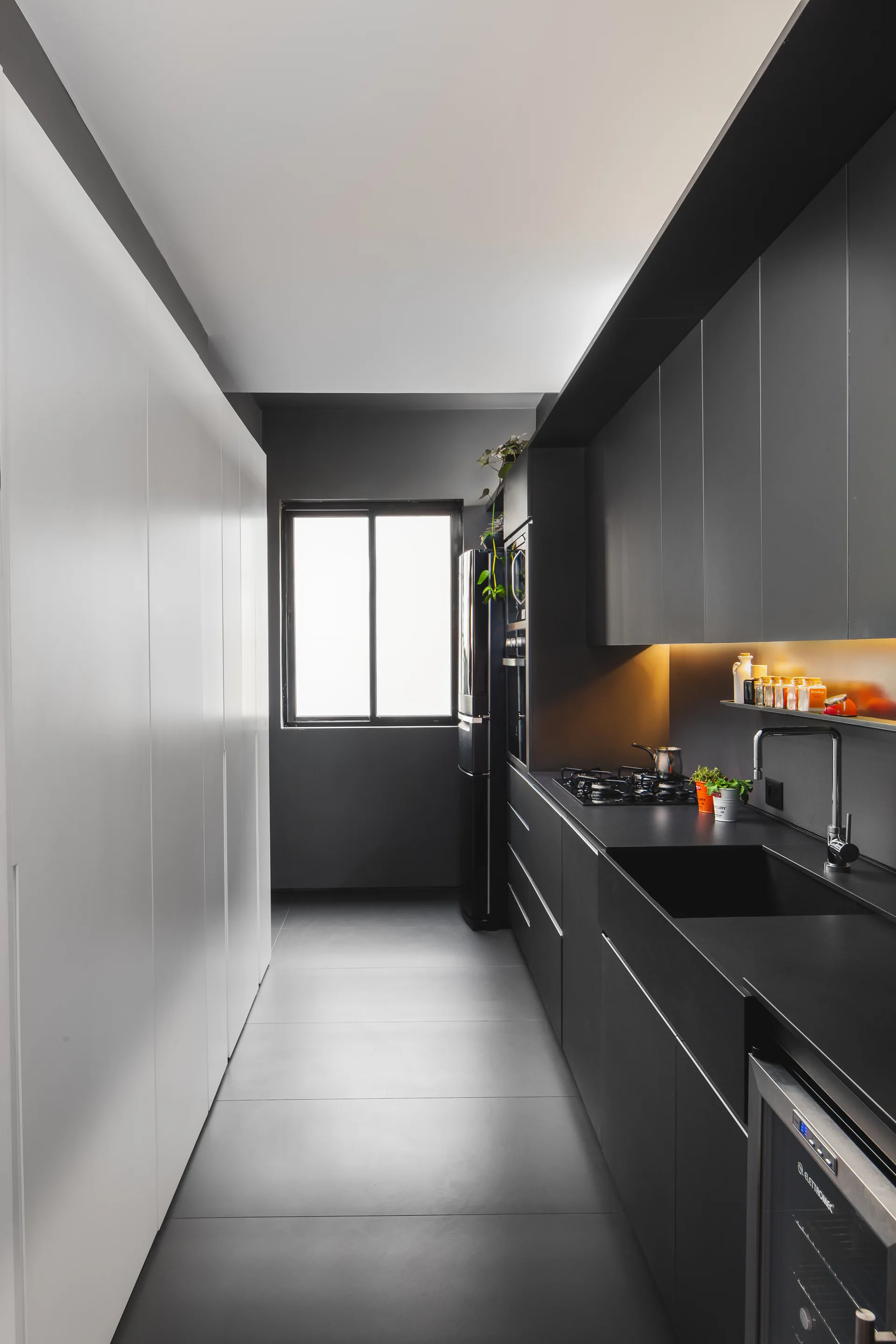 A narrow galley kitchen features matte black cabinets and a black sink on the right, contrasted by a floor-to-ceiling white wall of panels on the left.