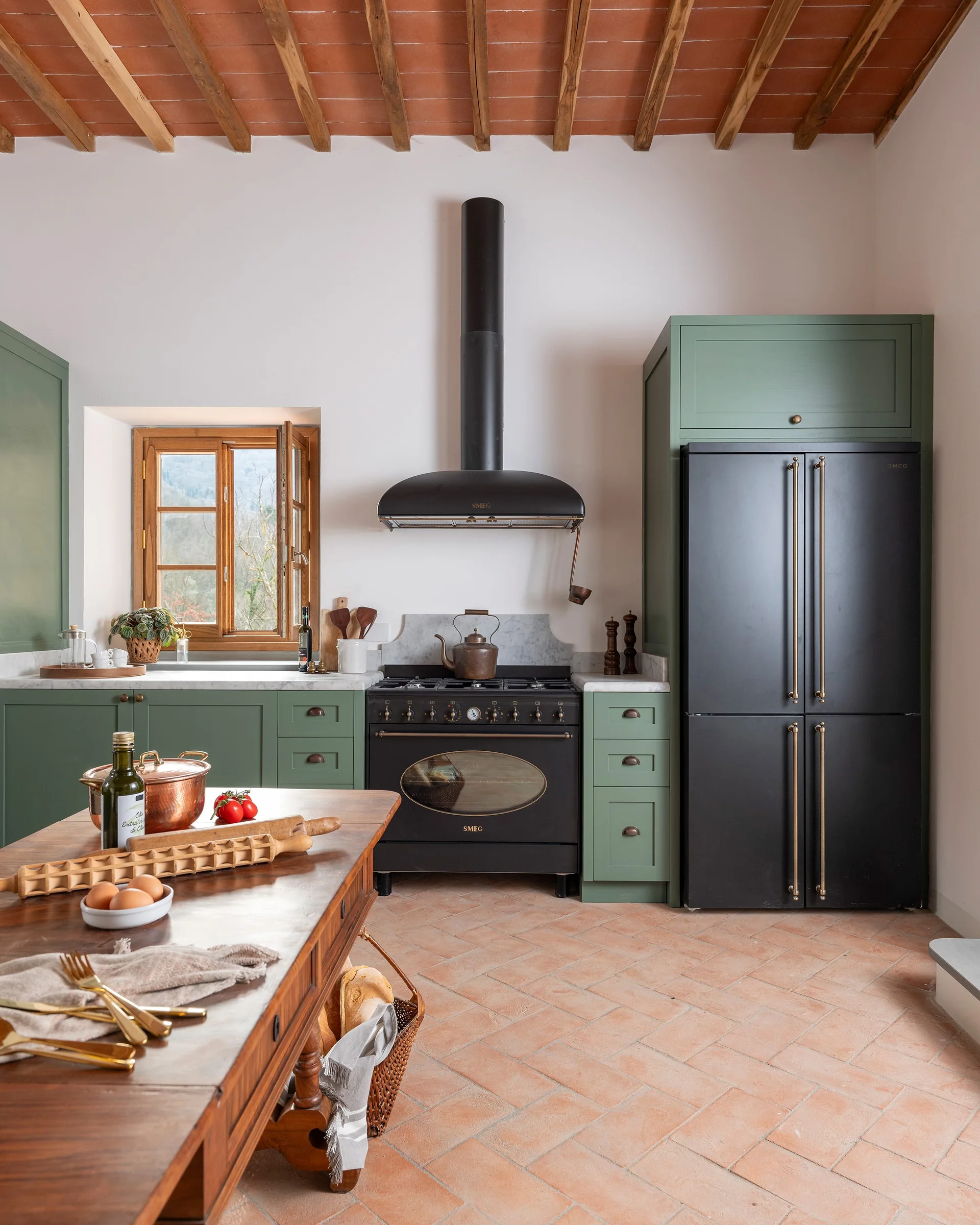 A farmhouse kitchen featuring sage green cabinets, a black stove and refrigerator, and terracotta tile flooring under exposed wood beams.