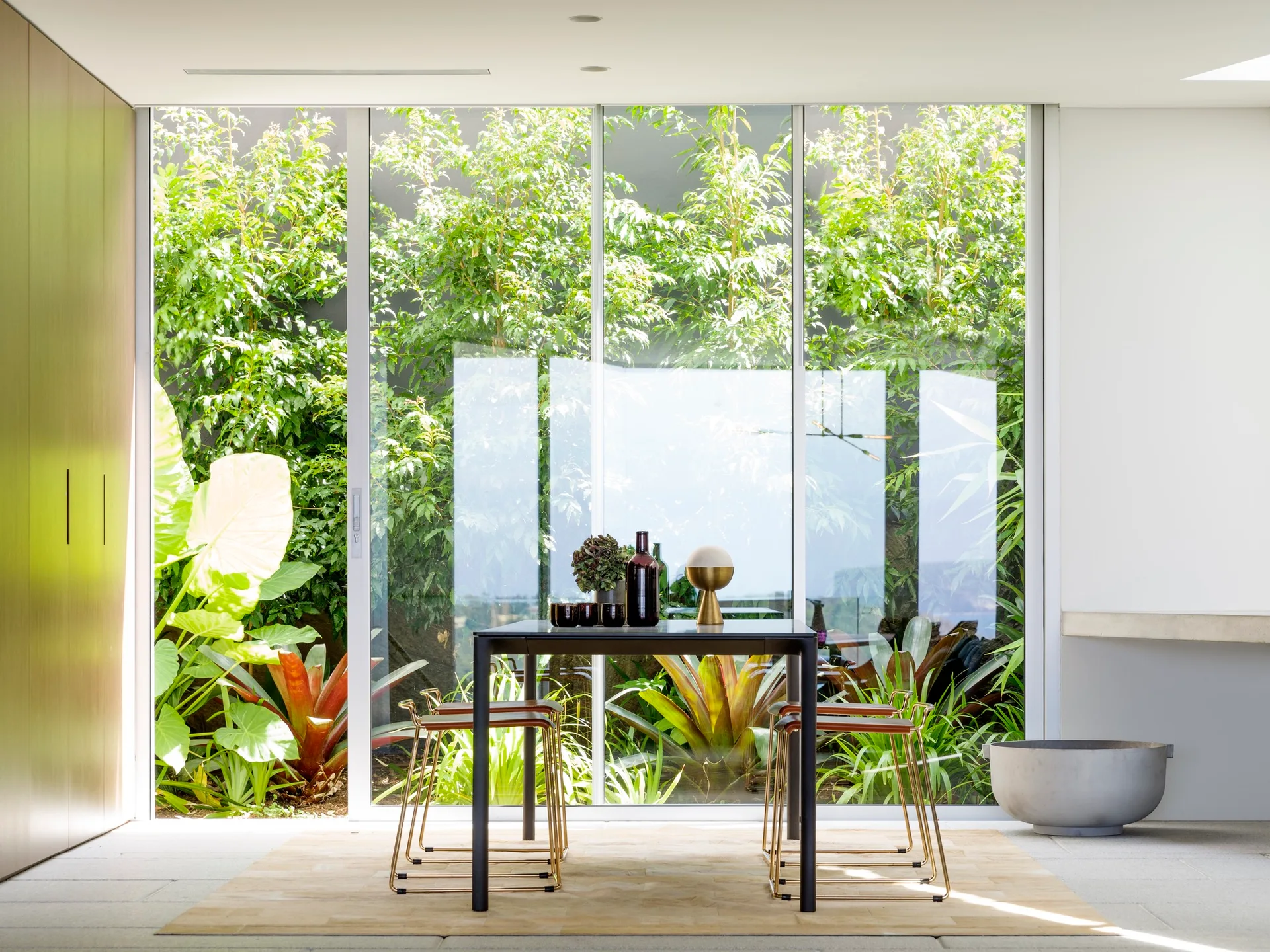 A dark dining table and brass-framed stools sit on a light rug in front of a floor-to-ceiling glass wall showcasing a lush green garden.