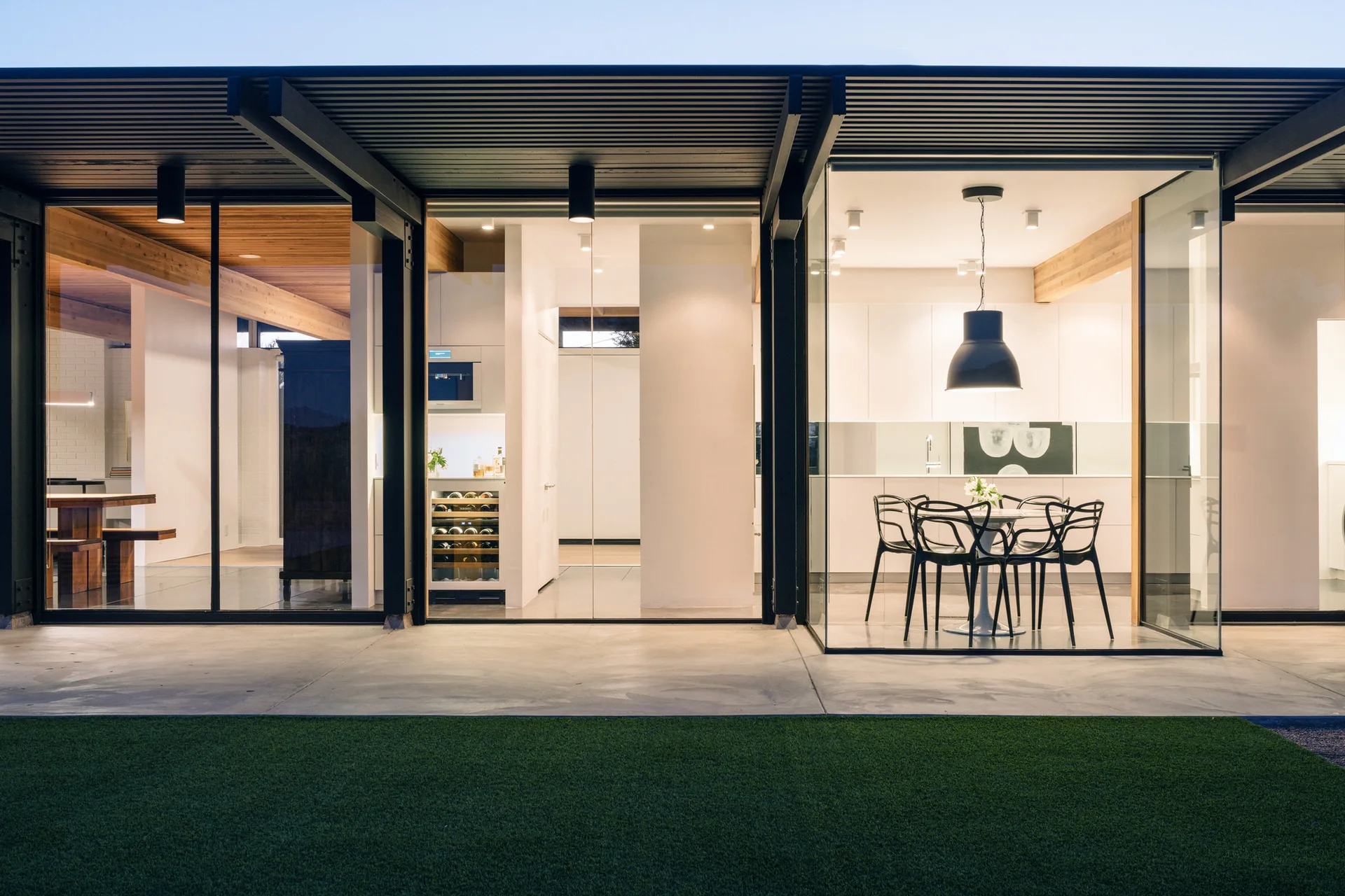 A modern home features an open-plan kitchen and dining area with white cabinetry, a built-in wine cooler, and black chairs visible through glass walls.
