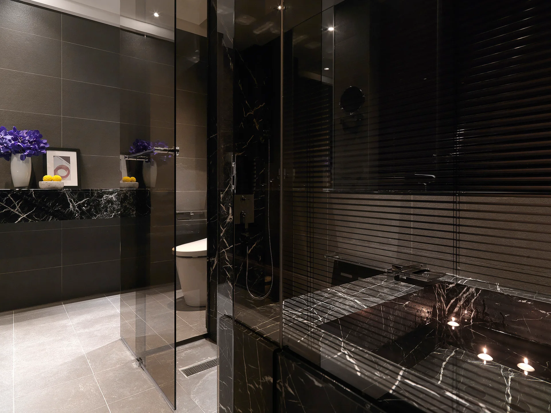 A modern dark bathroom features a black veined marble bathtub, a frameless glass shower panel, large grey floor tiles, and a white toilet.