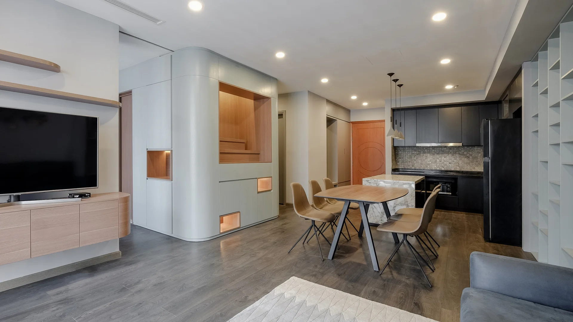 A modern open-plan interior features a curved light-grey room divider, a wooden dining table with four chairs, and a dark kitchen in the background.