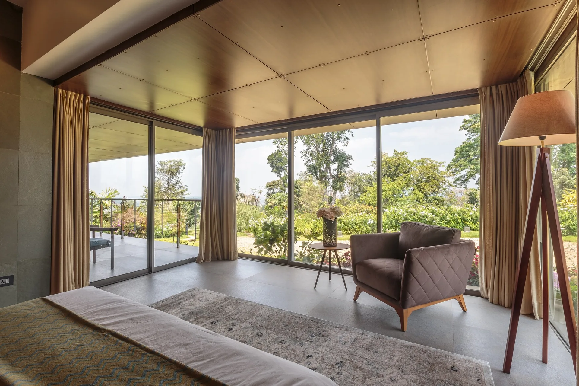 A bedroom sitting area features a brown quilted armchair, a wood tripod floor lamp, and a wood-paneled ceiling next to large sliding glass walls.