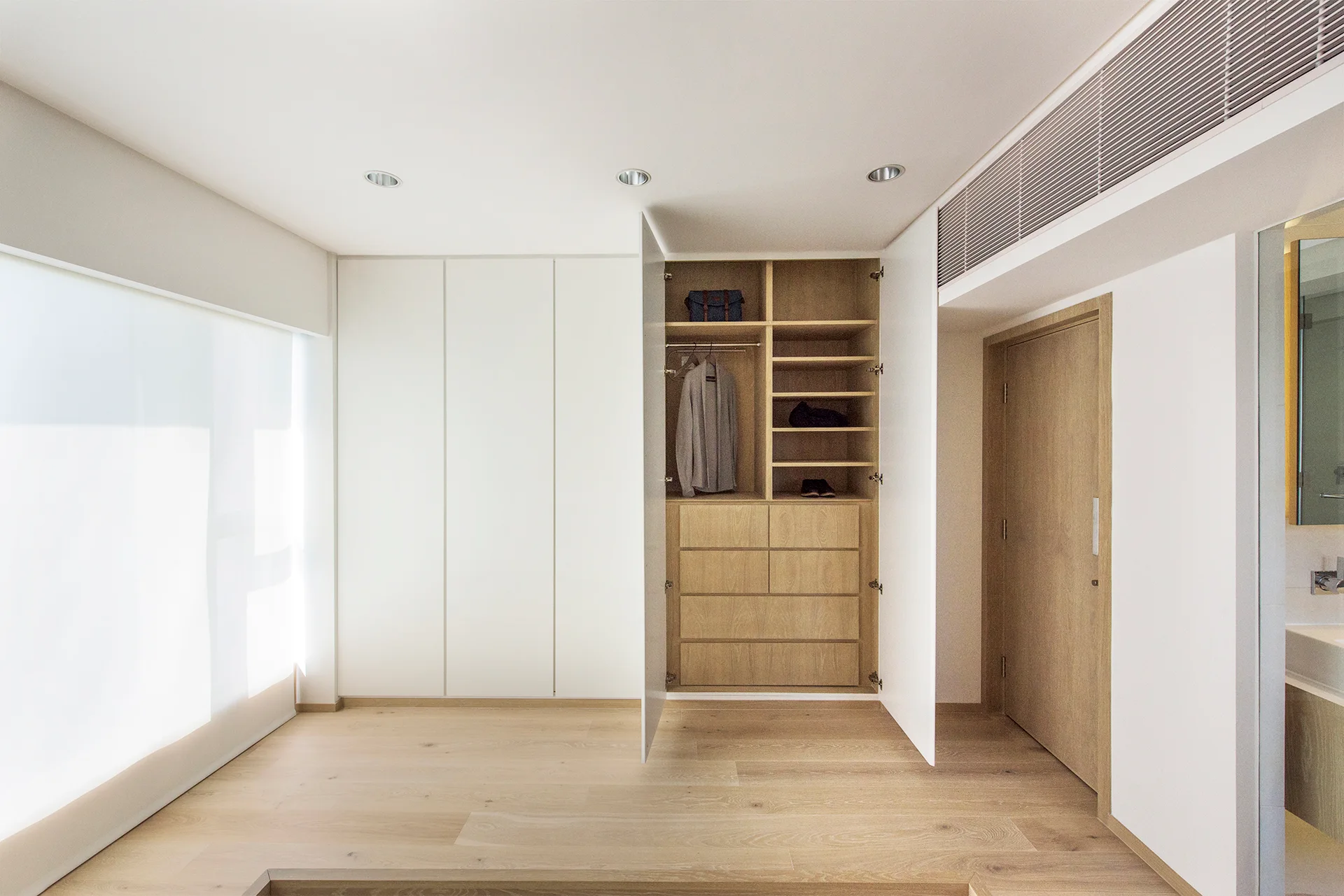 A minimalist room features a built-in white wardrobe with one open section showing a light wood interior, matching wood floors, and a wooden door.