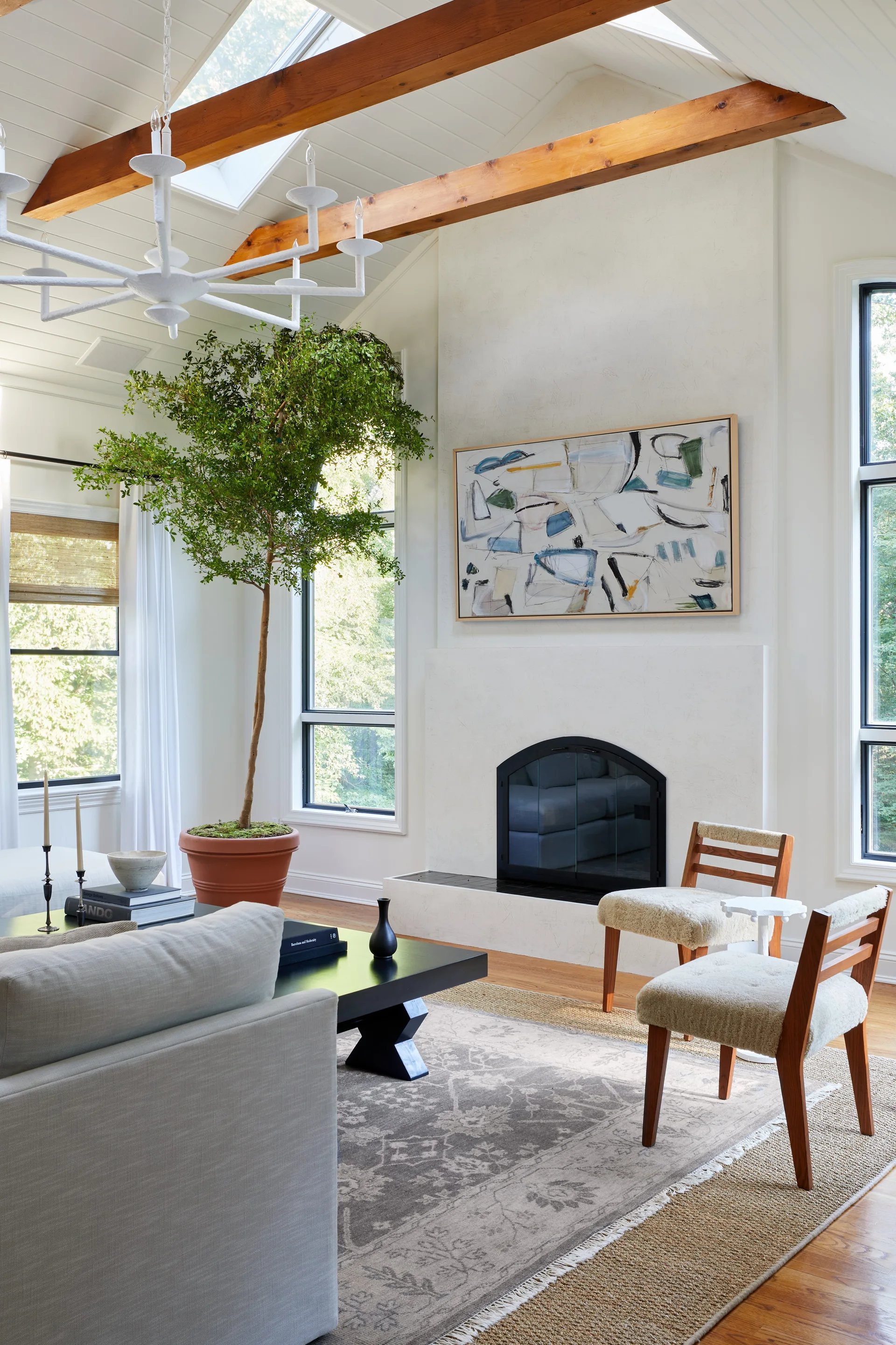 A bright living room features a white fireplace, exposed wood ceiling beams, a tall potted tree, and two accent chairs.