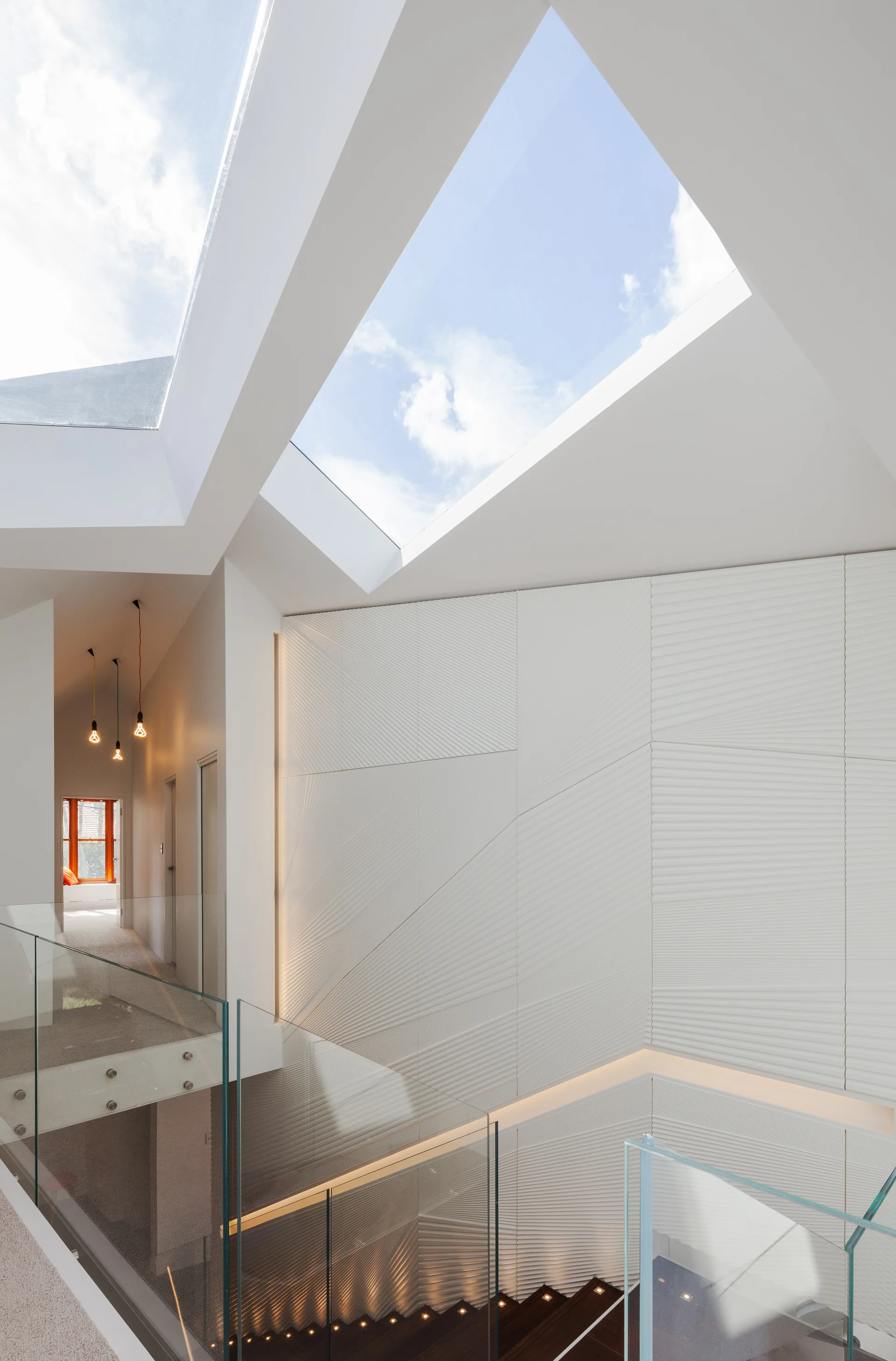 A modern staircase features geometric white textured wall panels illuminated by large ceiling skylights next to a glass railing.