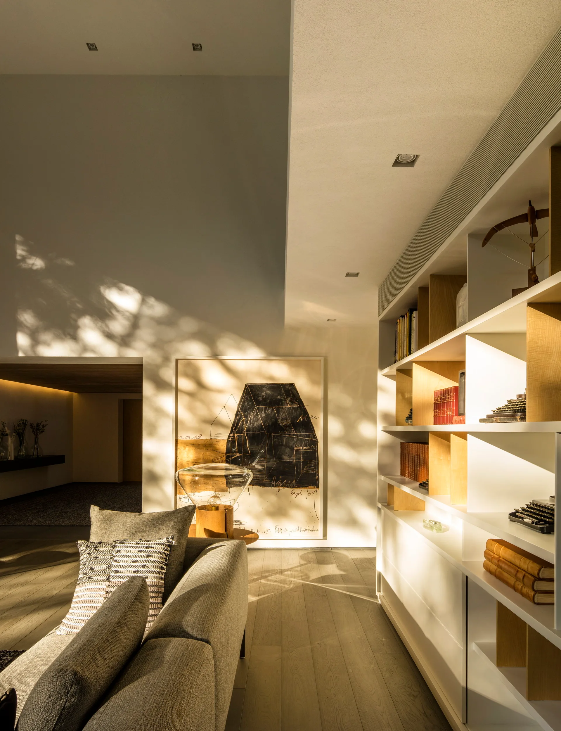 A gray fabric sofa stands near a tall white and wood built-in bookshelf and a large abstract painting on a sunlit wall.