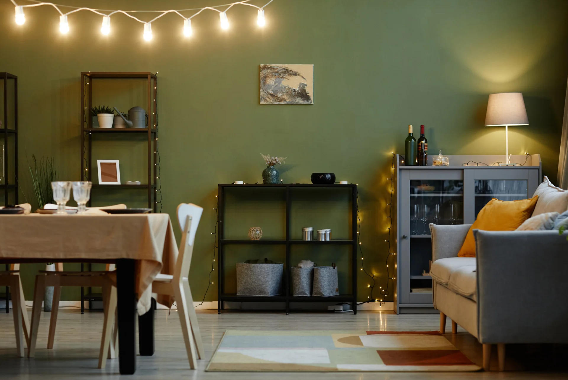 A living and dining space features an olive green wall, a grey sofa with a mustard pillow, string lights, and a table with a beige tablecloth.