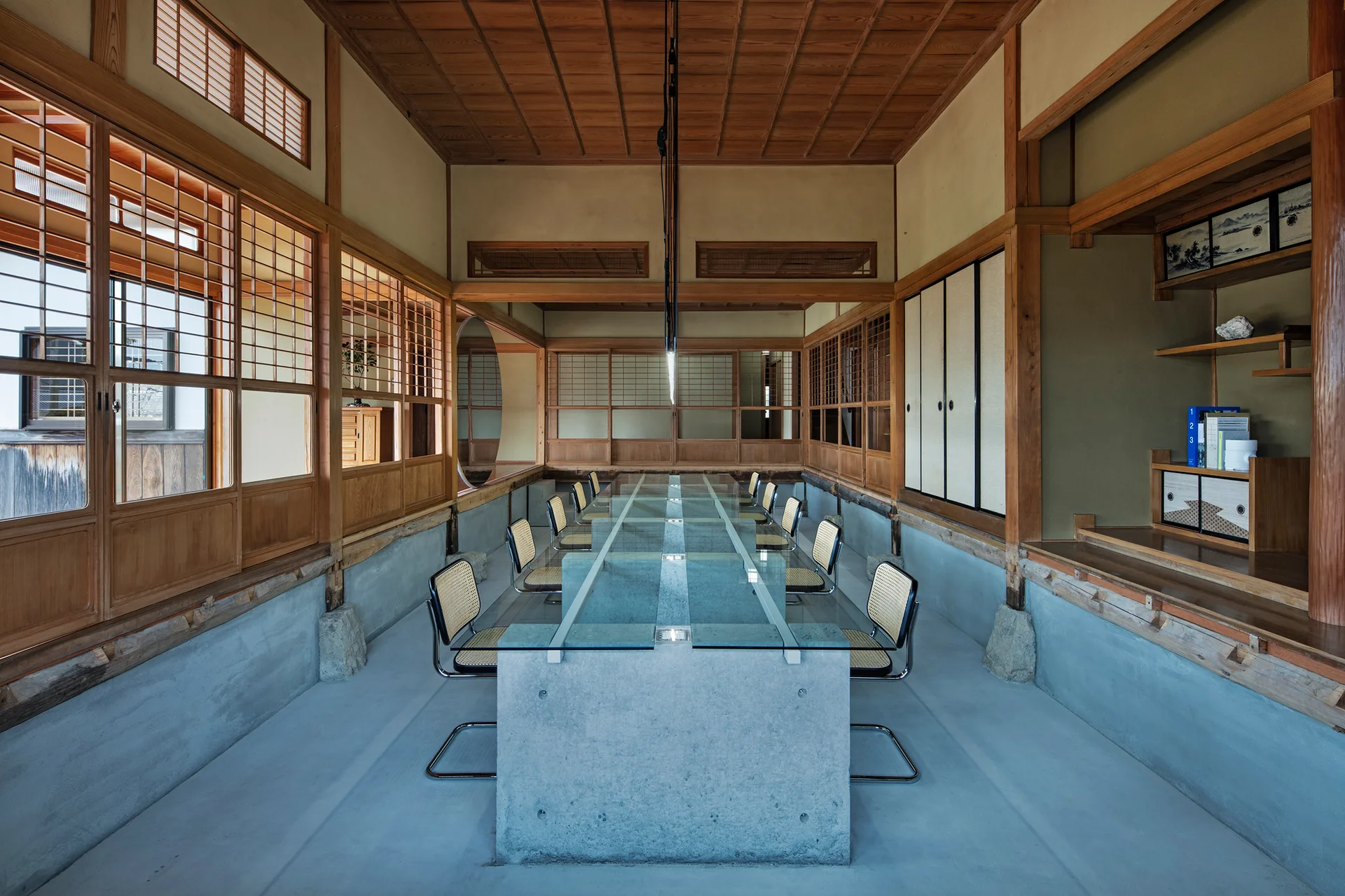 A long glass and concrete dining table surrounded by cane chairs inside a traditional Japanese room with wood paneled walls and shoji screens.