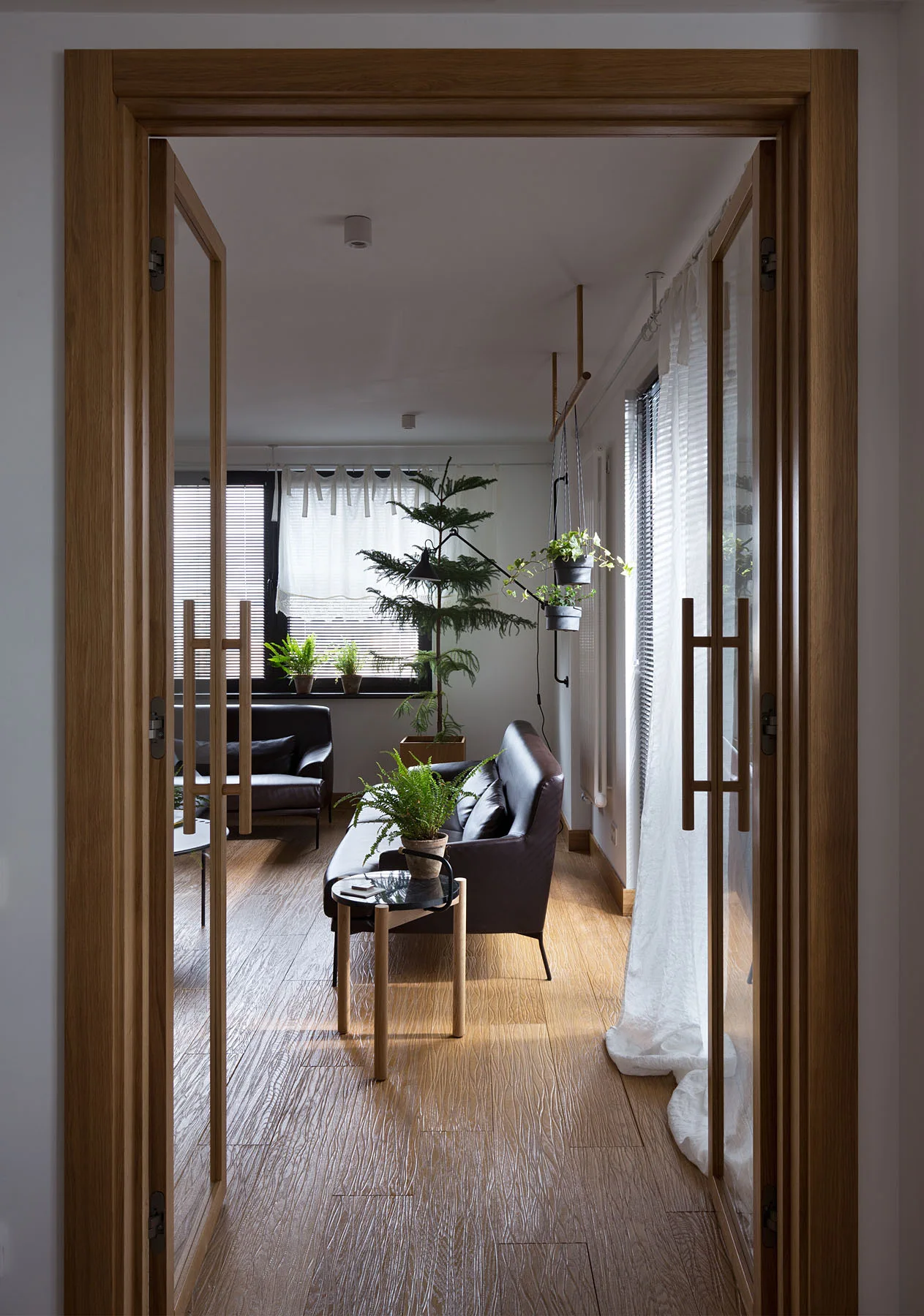 A view through a wooden-framed glass doorway into a living room with dark leather sofas, sheer white curtains, and multiple potted houseplants.