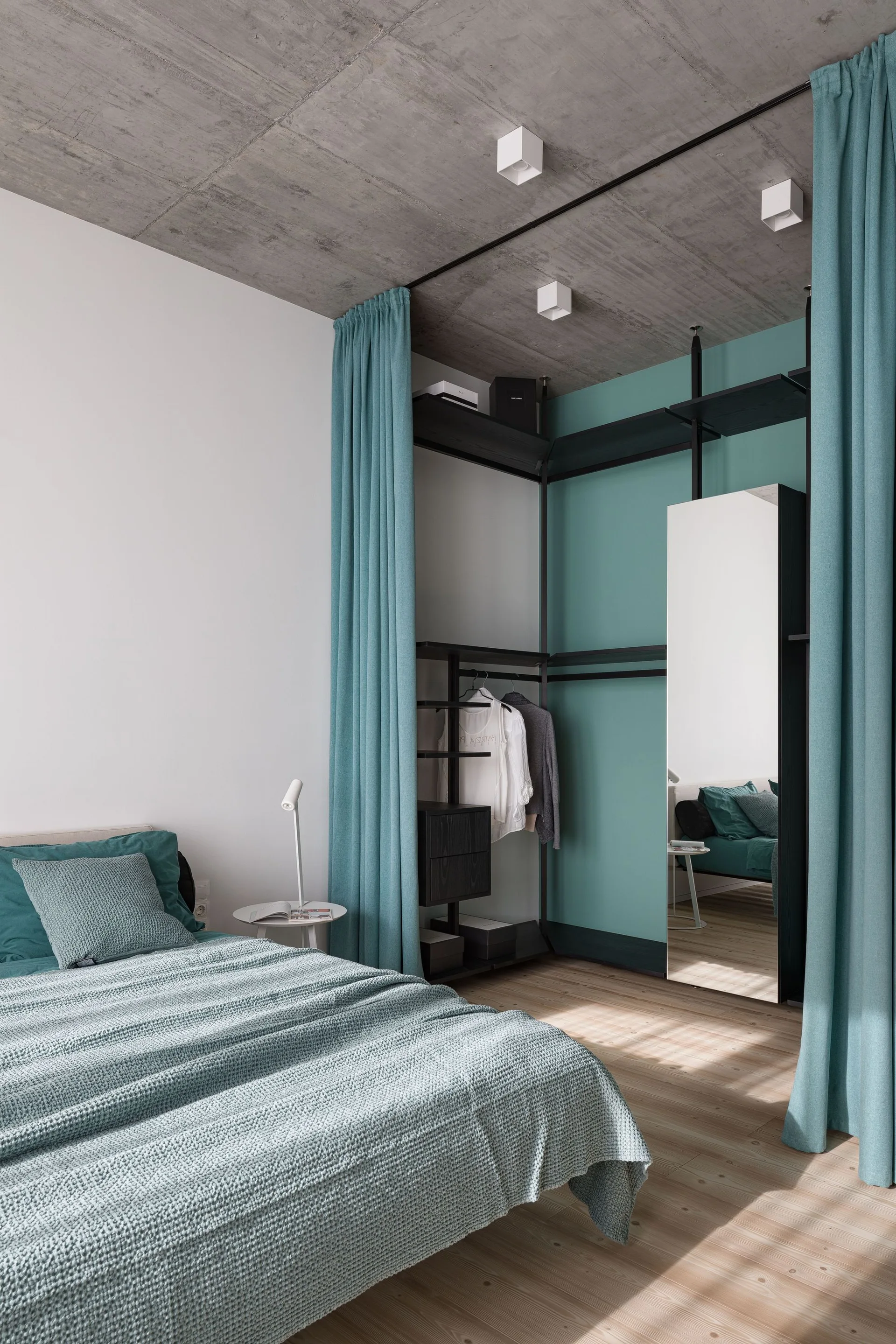 A modern bedroom features an exposed concrete ceiling, a bed with teal bedding, and a black open-frame wardrobe enclosed by tall teal curtains.