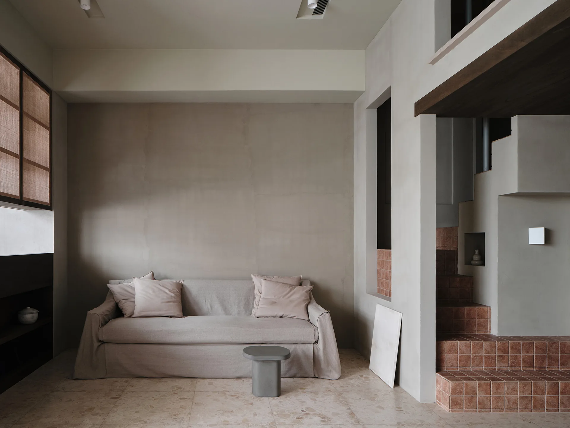 A minimalist living room features a beige slipcovered sofa against a plaster wall, next to a staircase covered in square terracotta tiles.