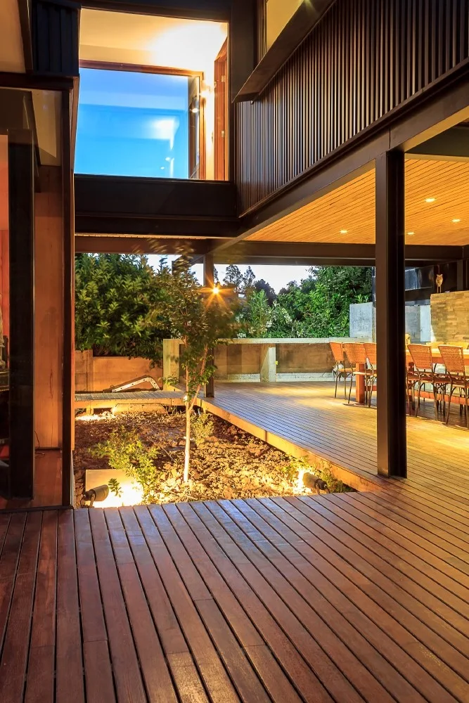 A wooden outdoor deck at dusk with a sunken garden bed, a small tree, and an outdoor dining set under a wood-clad ceiling.