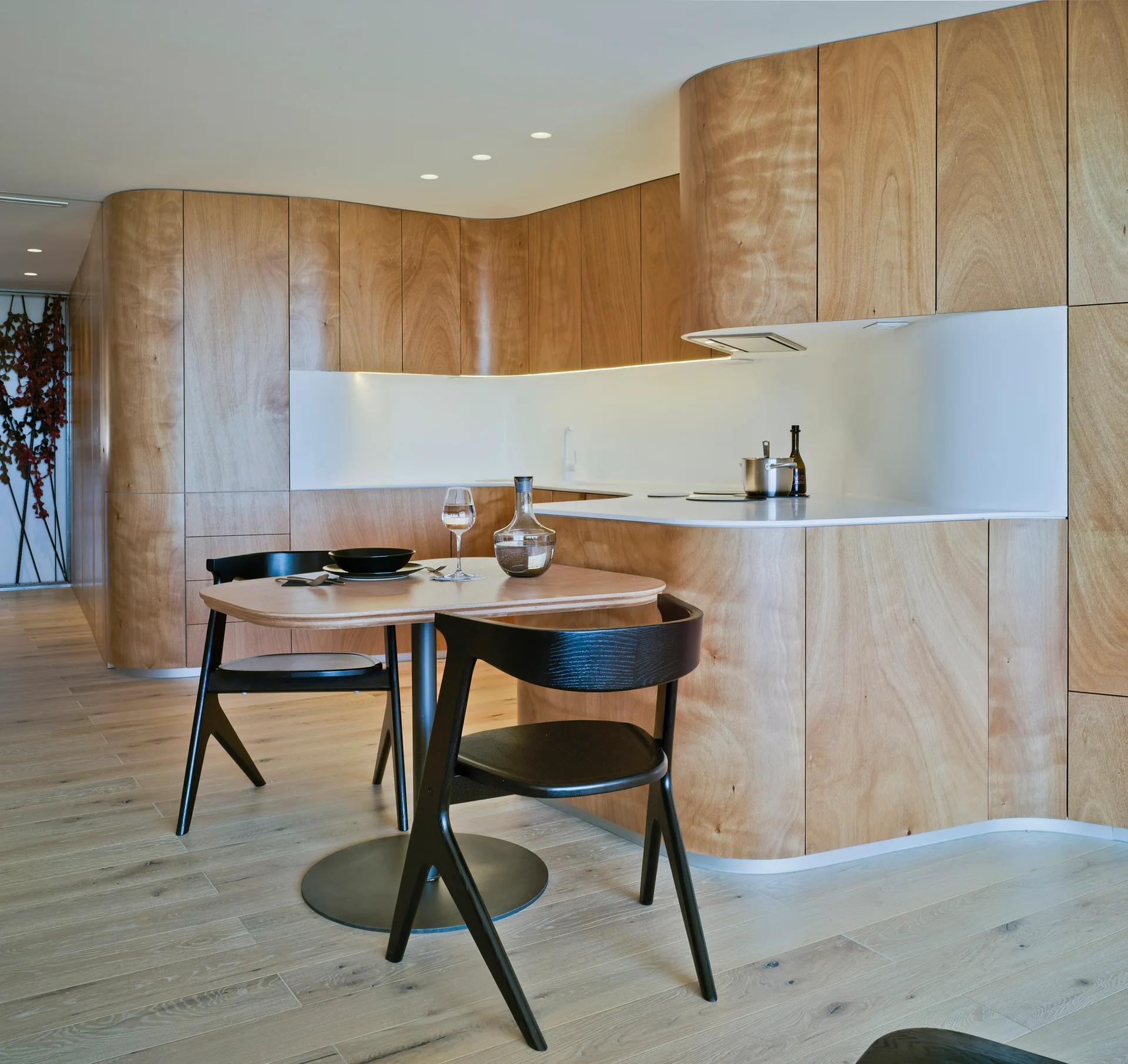 A modern kitchen features curved wood cabinetry, under-cabinet lighting, a white backsplash, and a small wood dining table with two black chairs.