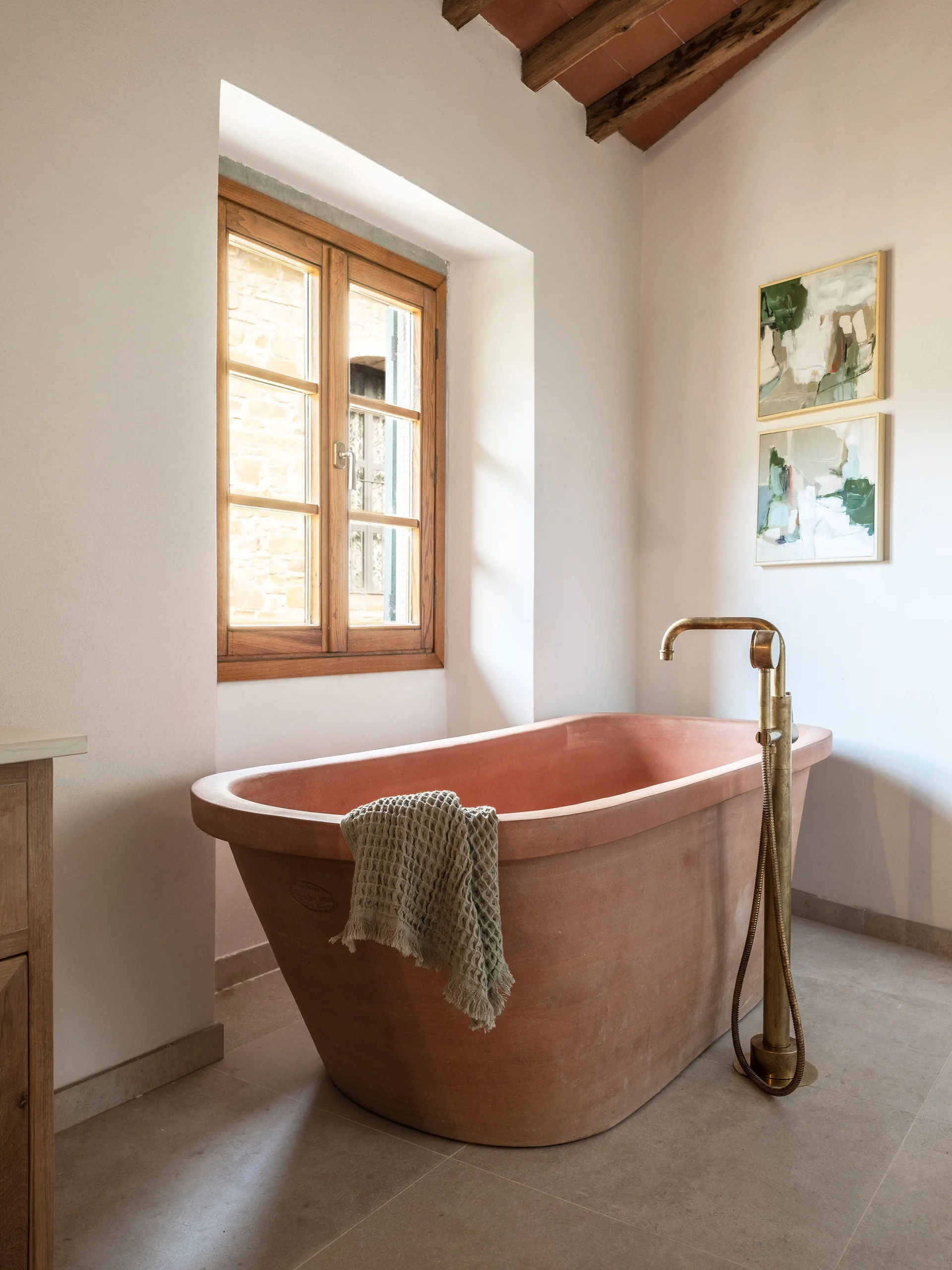 A terracotta freestanding bathtub sits below a wooden window in a rustic bathroom with plaster walls, a brass faucet, and exposed ceiling beams.