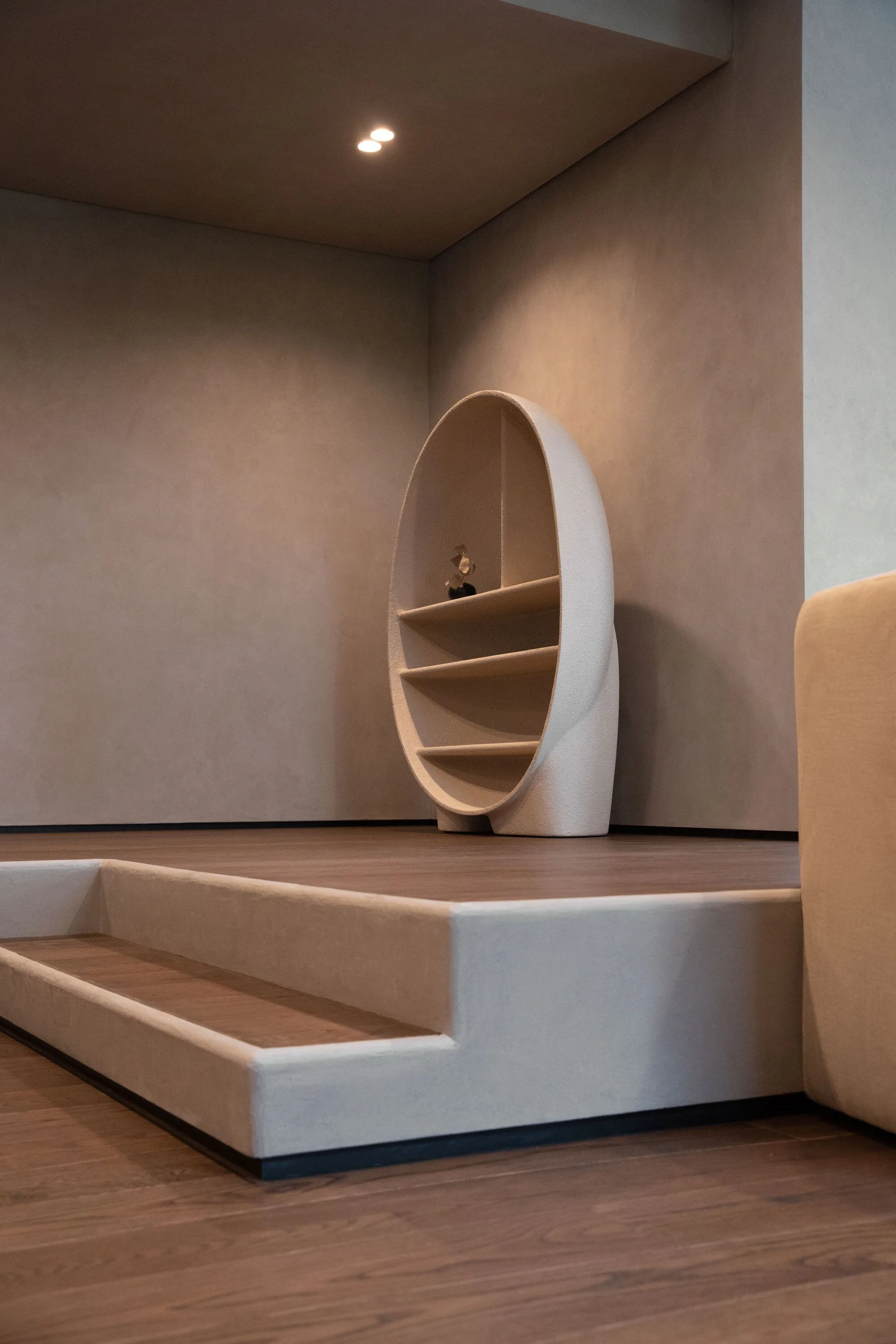 A minimalist oval shelving unit stands on a raised wooden platform with steps in a warm beige plastered room.