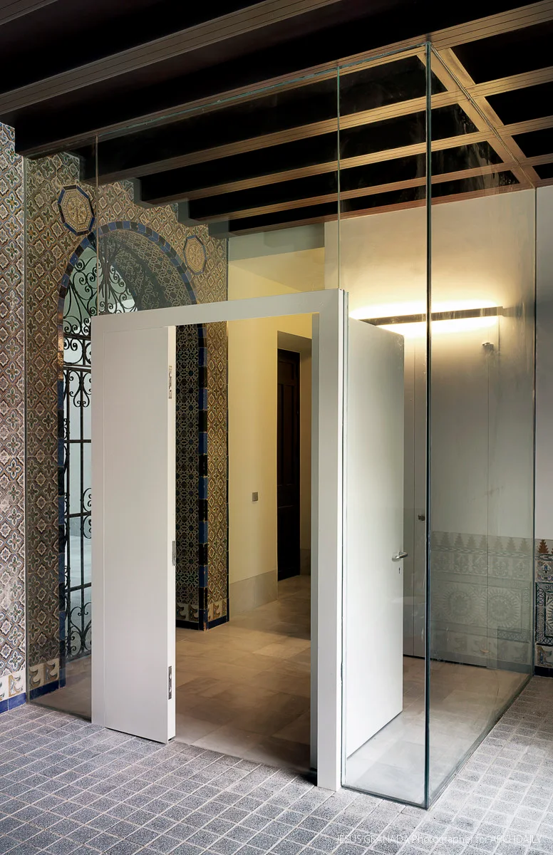 A minimalist glass enclosure with a white door stands inside a room featuring ornate patterned wall tiles and a dark wood grid ceiling.