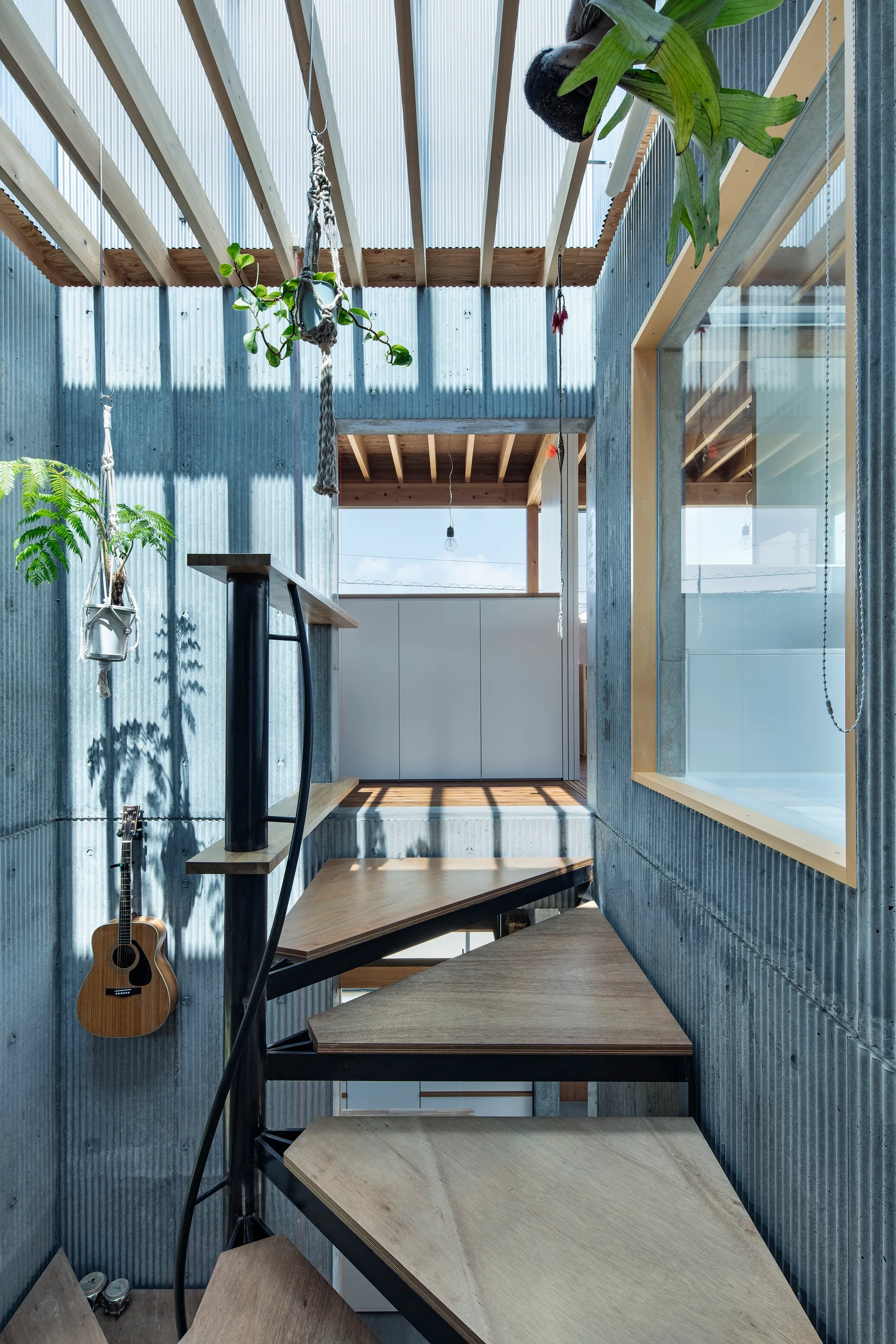 A metal spiral staircase with wood treads rises next to a grey corrugated metal wall under a translucent ceiling with exposed wooden rafters.
