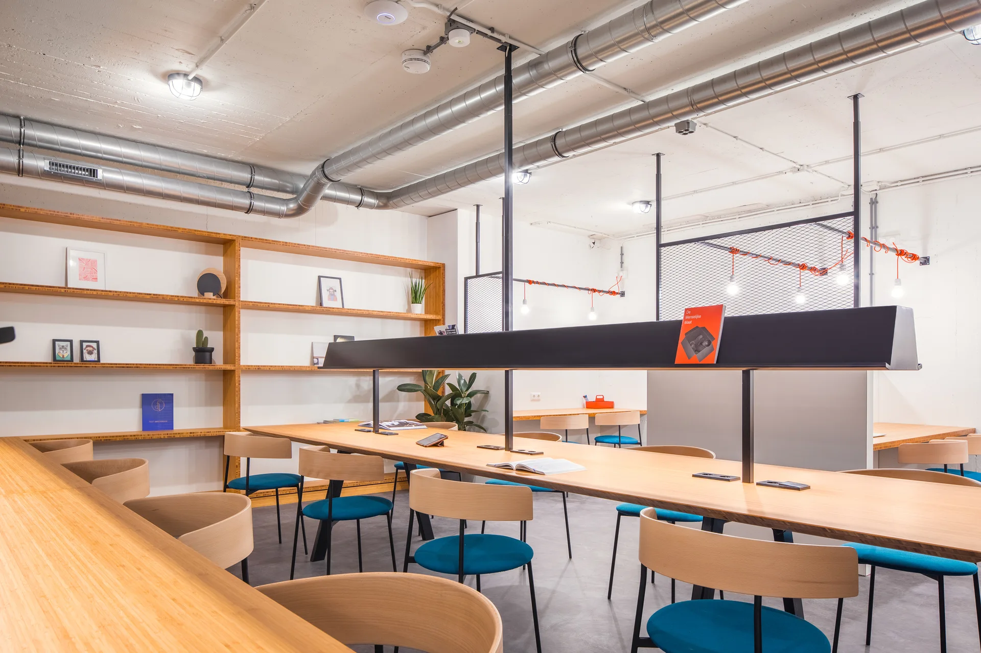A modern open-plan workspace featuring a long wooden desk with blue-cushioned chairs, light wood wall shelves, and exposed ceiling ductwork.
