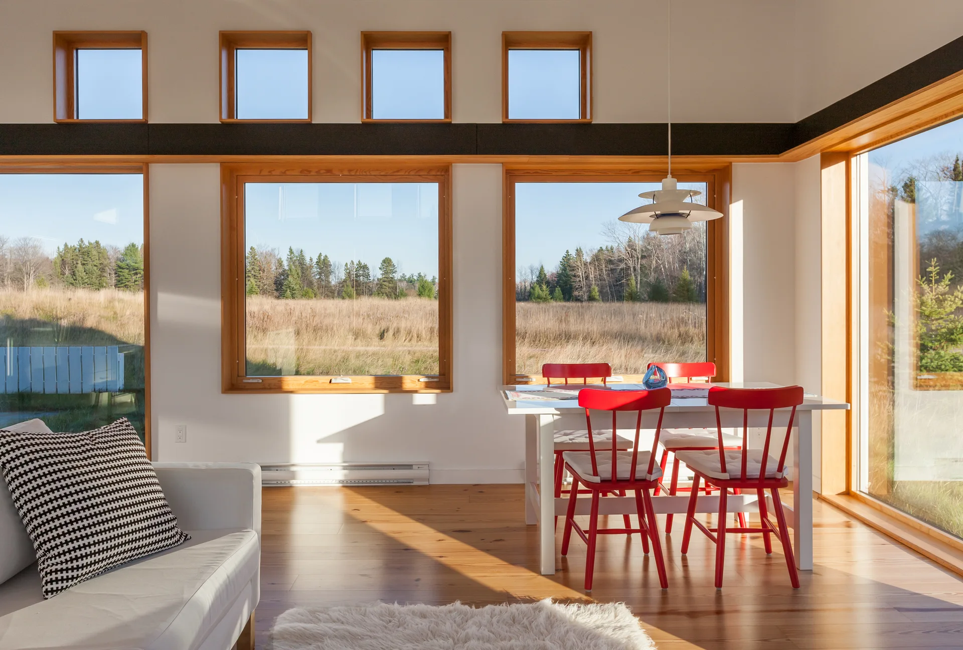 A bright dining area featuring a white table with four red wooden chairs next to large wood-framed windows and a row of high square windows.