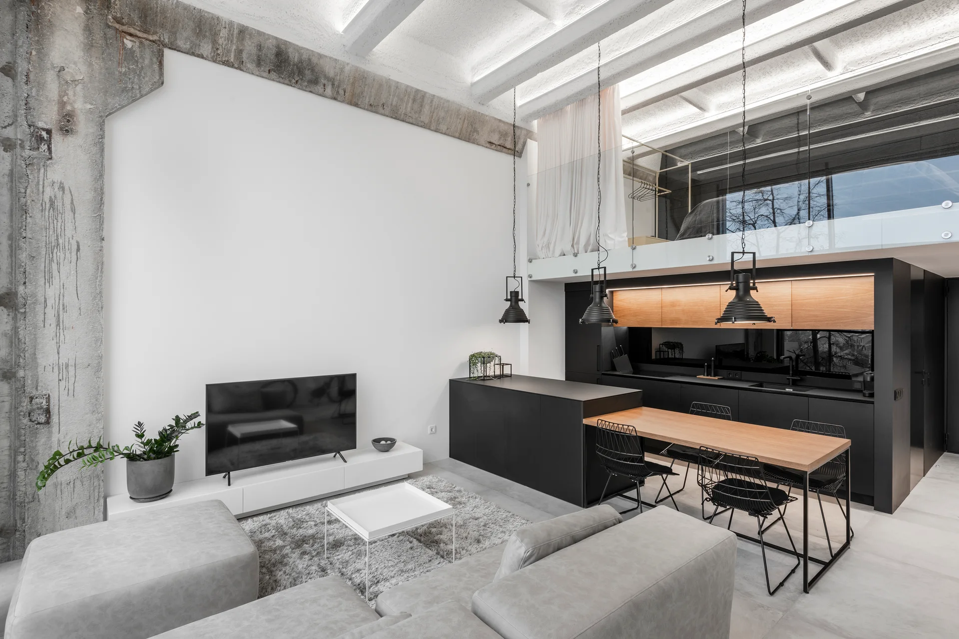 A modern loft living room features a grey sectional sofa, a matte black kitchen under a mezzanine, and an exposed concrete column.