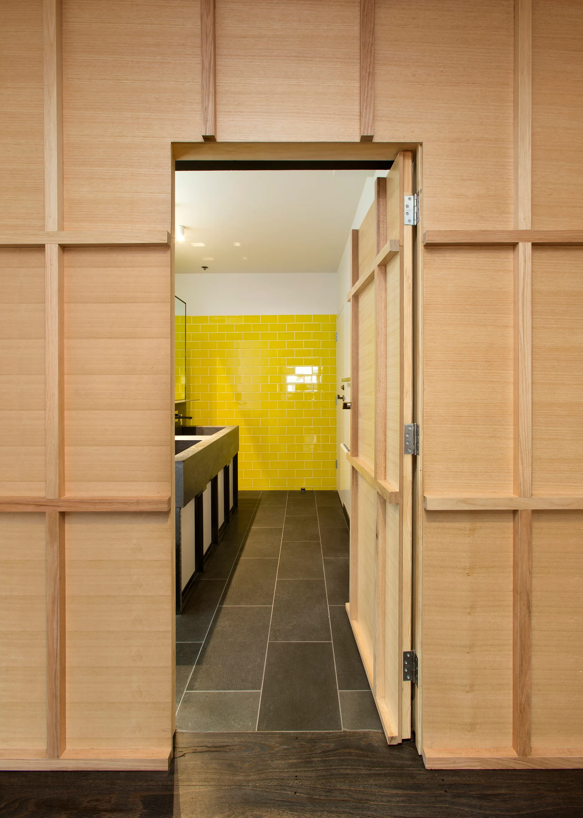 A view through an open light wood paneled doorway into a modern bathroom featuring a bright yellow subway tile wall and dark grey stone floor tiles.