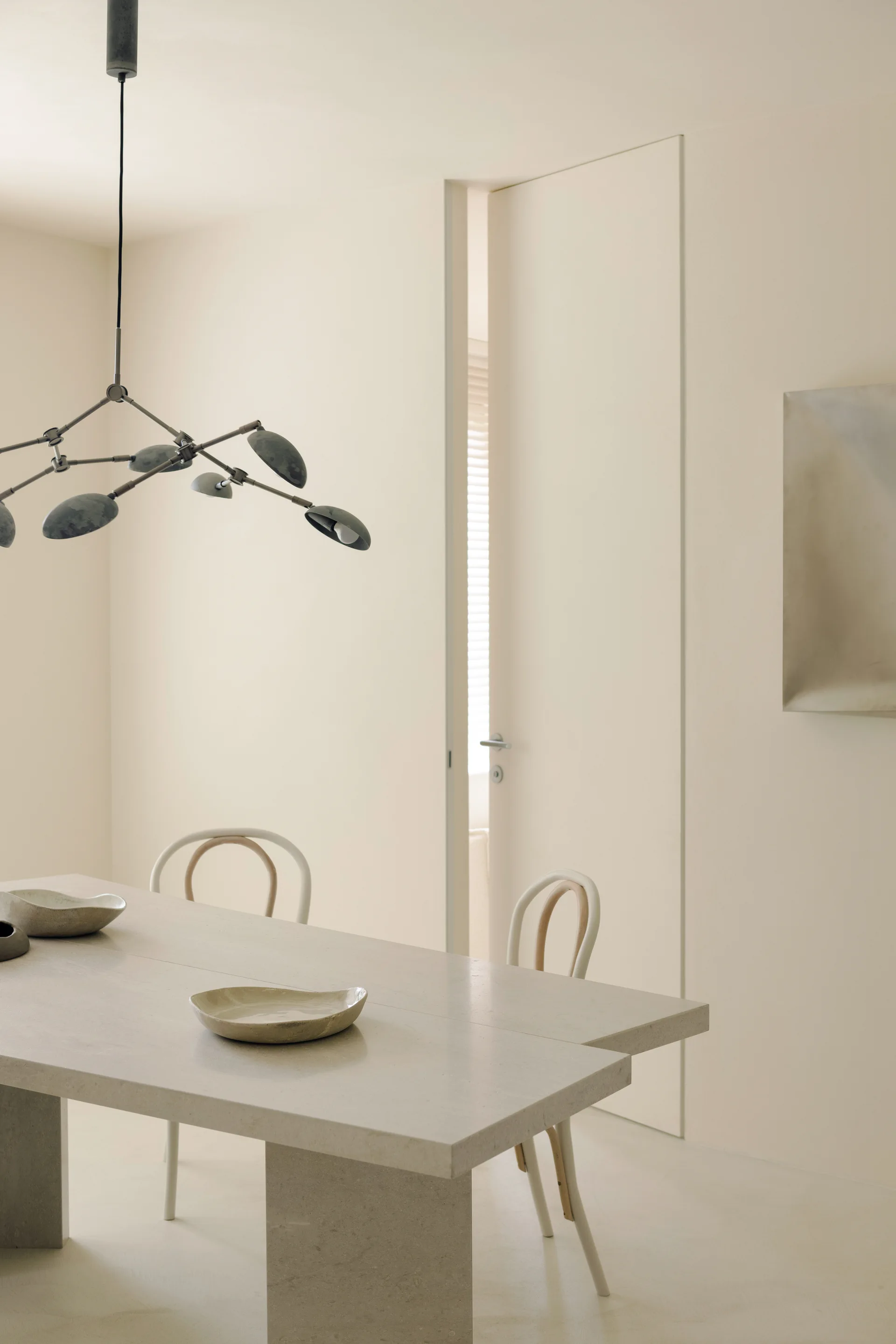 A minimalist cream dining room features a textured stone table with organic bowls, bicolored bentwood chairs, and a dark branching pendant light.