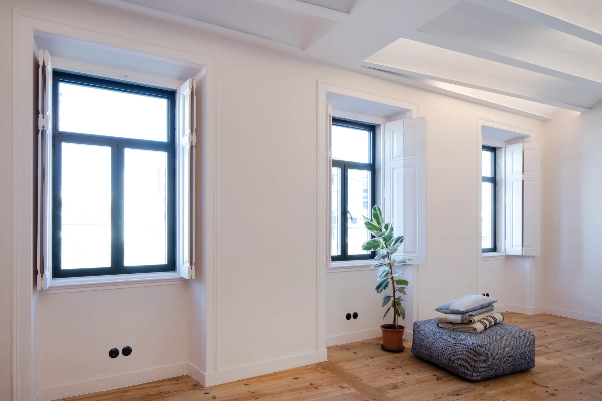 A sunlit minimalist room features three windows with white folding wooden shutters, light wood plank floors, and a blue patterned ottoman.