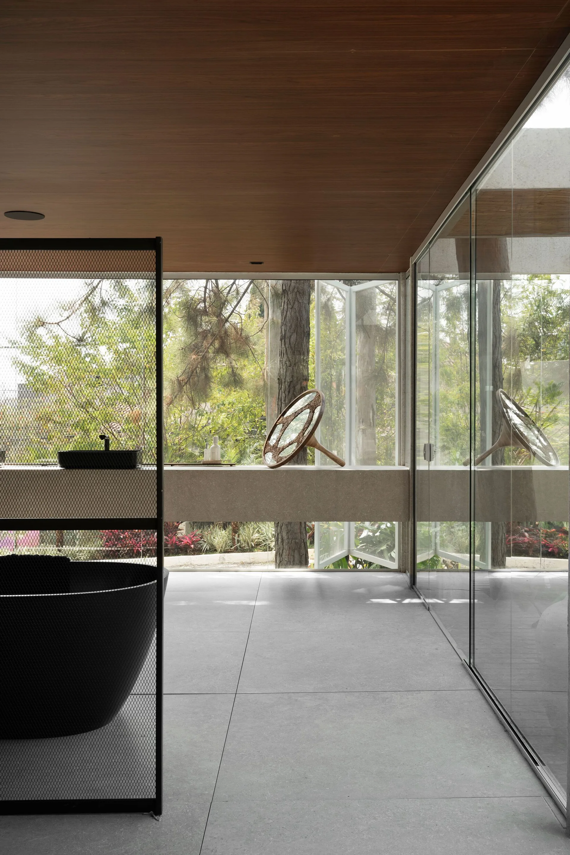 A modern bathroom features a black soaking tub, a floating stone vanity, and a wood ceiling next to a glass wall showing trees.
