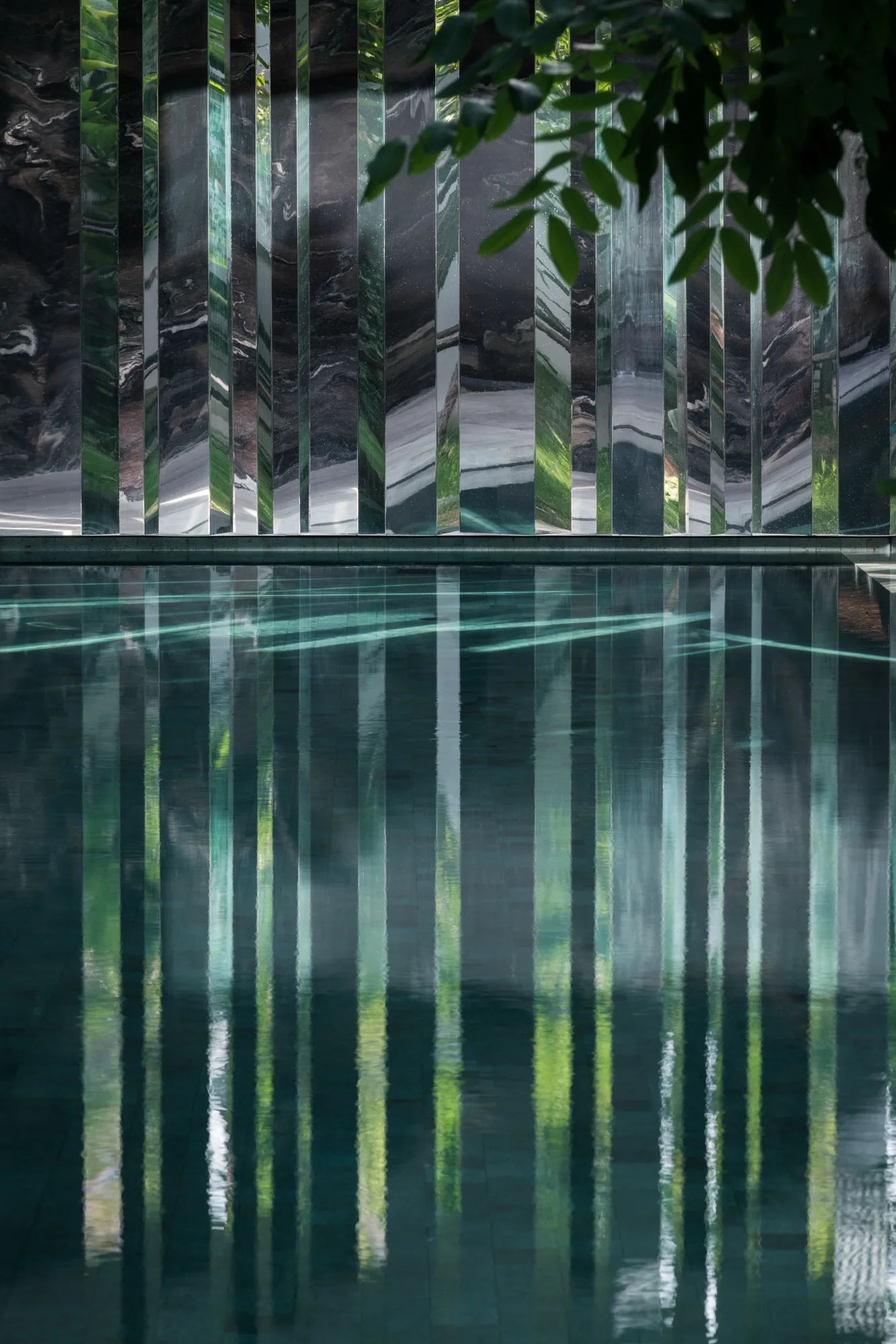 Still water in a modern outdoor pool reflects tall vertical mirrored panels and green leaves from above.
