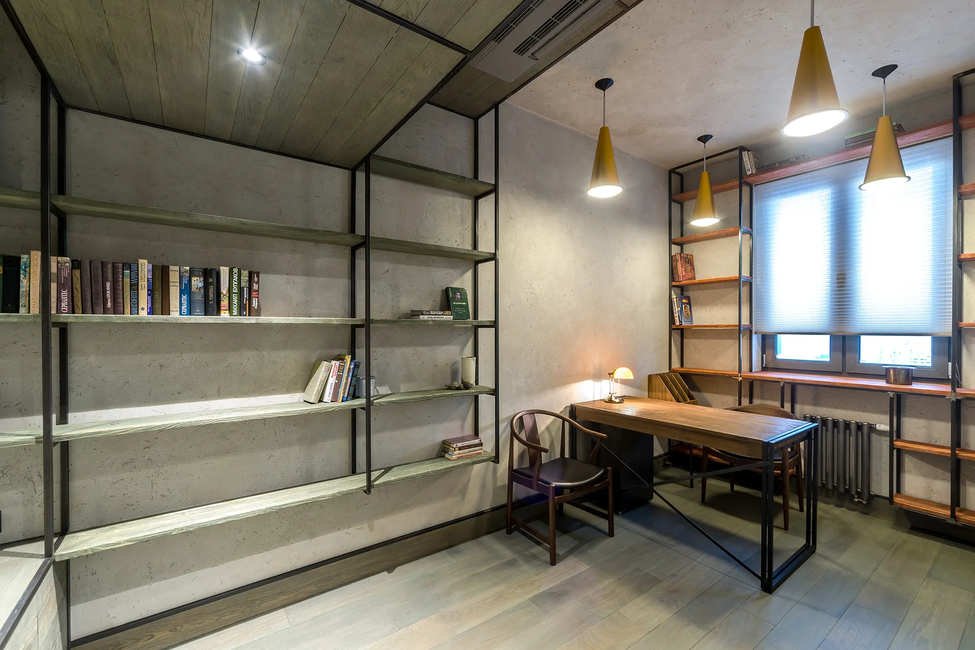 An industrial workspace featuring textured concrete walls, open black metal bookshelves, a wooden desk, and conical yellow pendant lights.